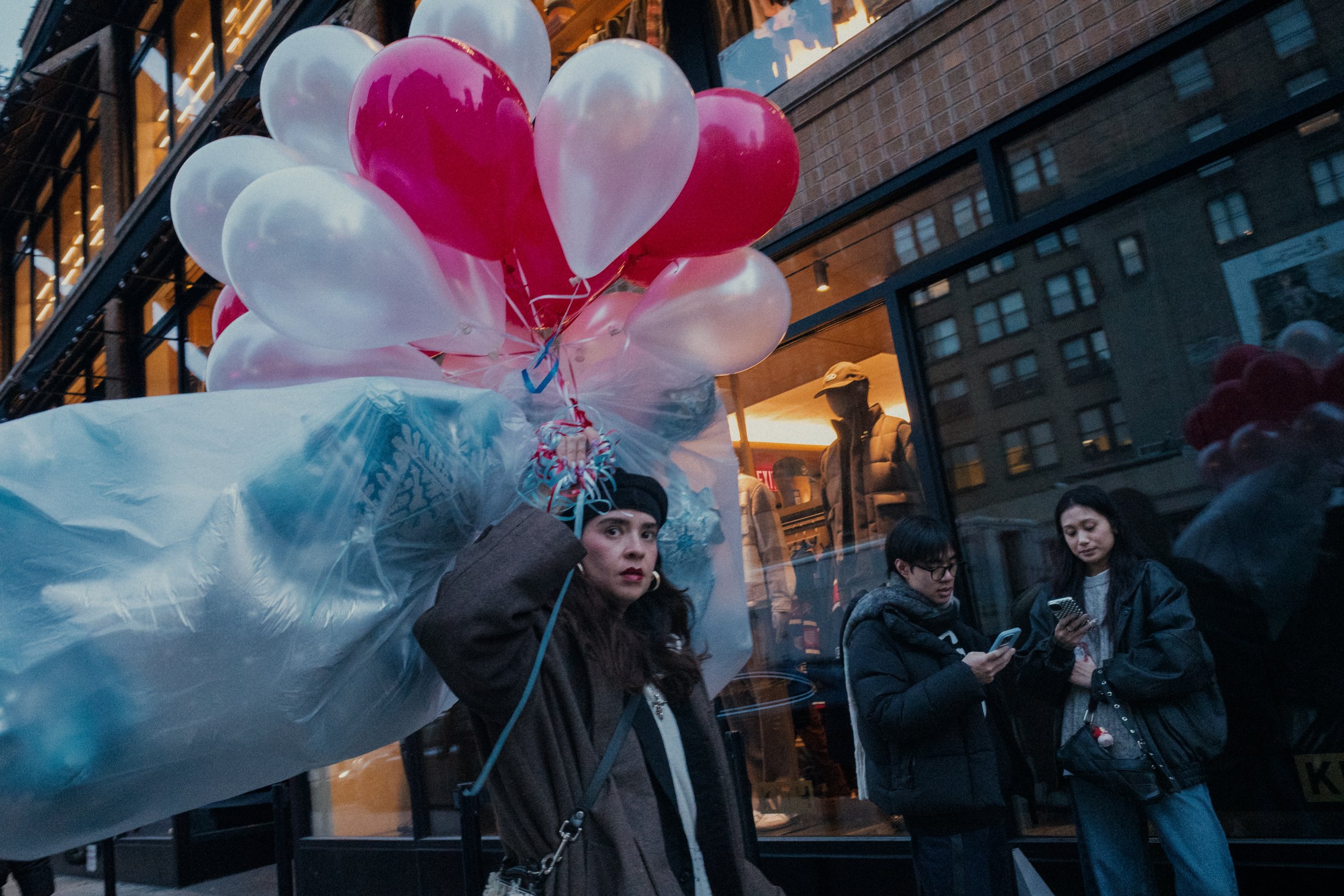 A woman holding a large cluster of pink and white balloons on a city street near a store window, with two people looking at their phones and a mannequin inside the store.