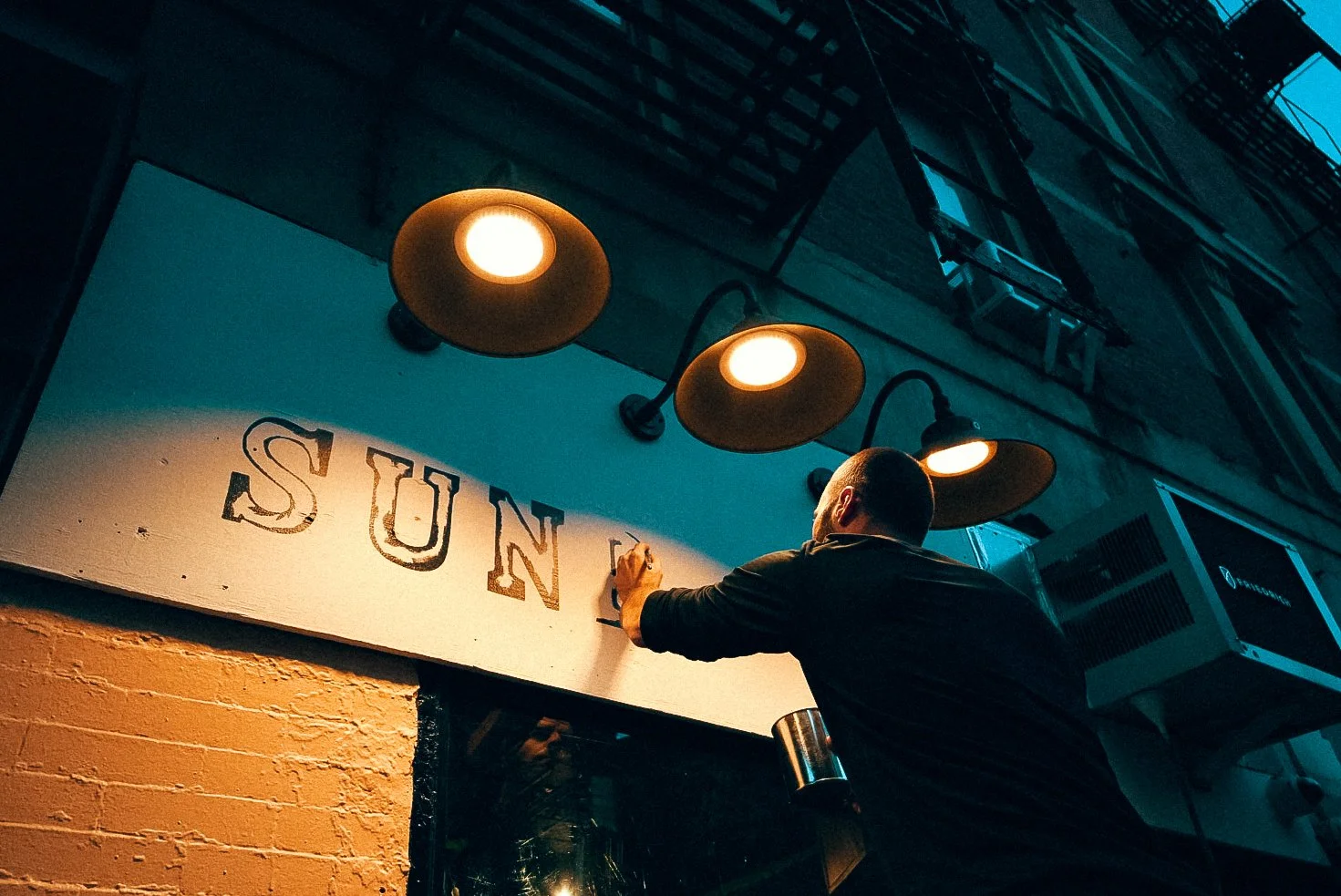 A man writes the word 'SUN' on a sign outside a restaurant or cafe under warm overhead lighting at dusk.
