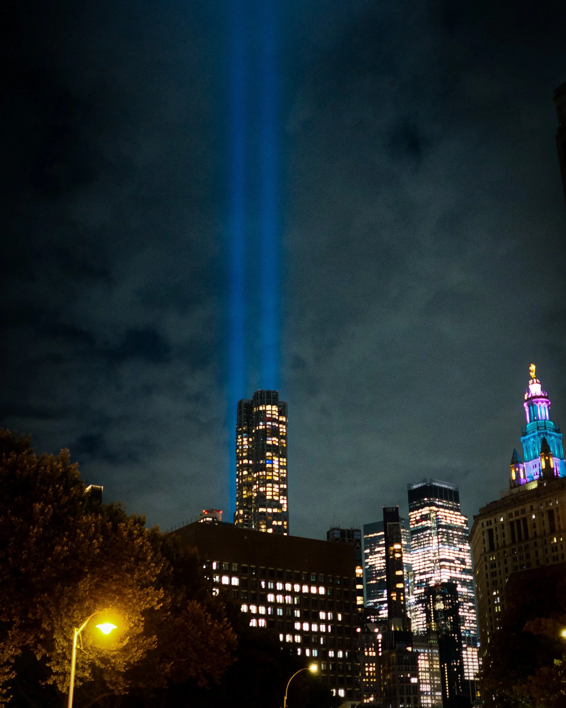 Nighttime cityscape with illuminated skyscrapers, including one with blue and purple lighting, and two blue beams of light projected into the sky.