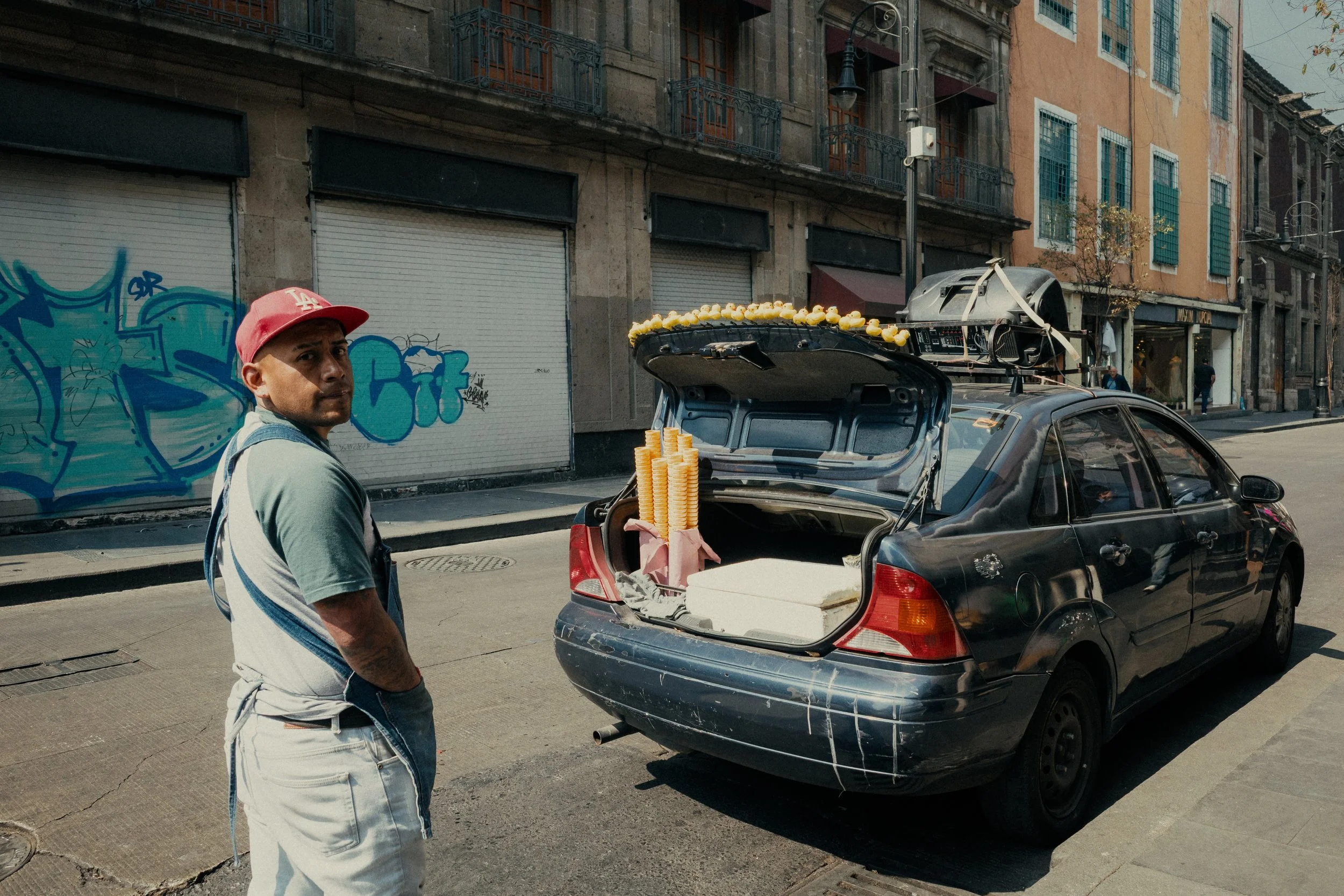 A man with a red cap and apron standing on a city street next to a black car with its trunk open, displaying stacks of yellow candies and white boxes, with graffiti-covered storefronts in the background.