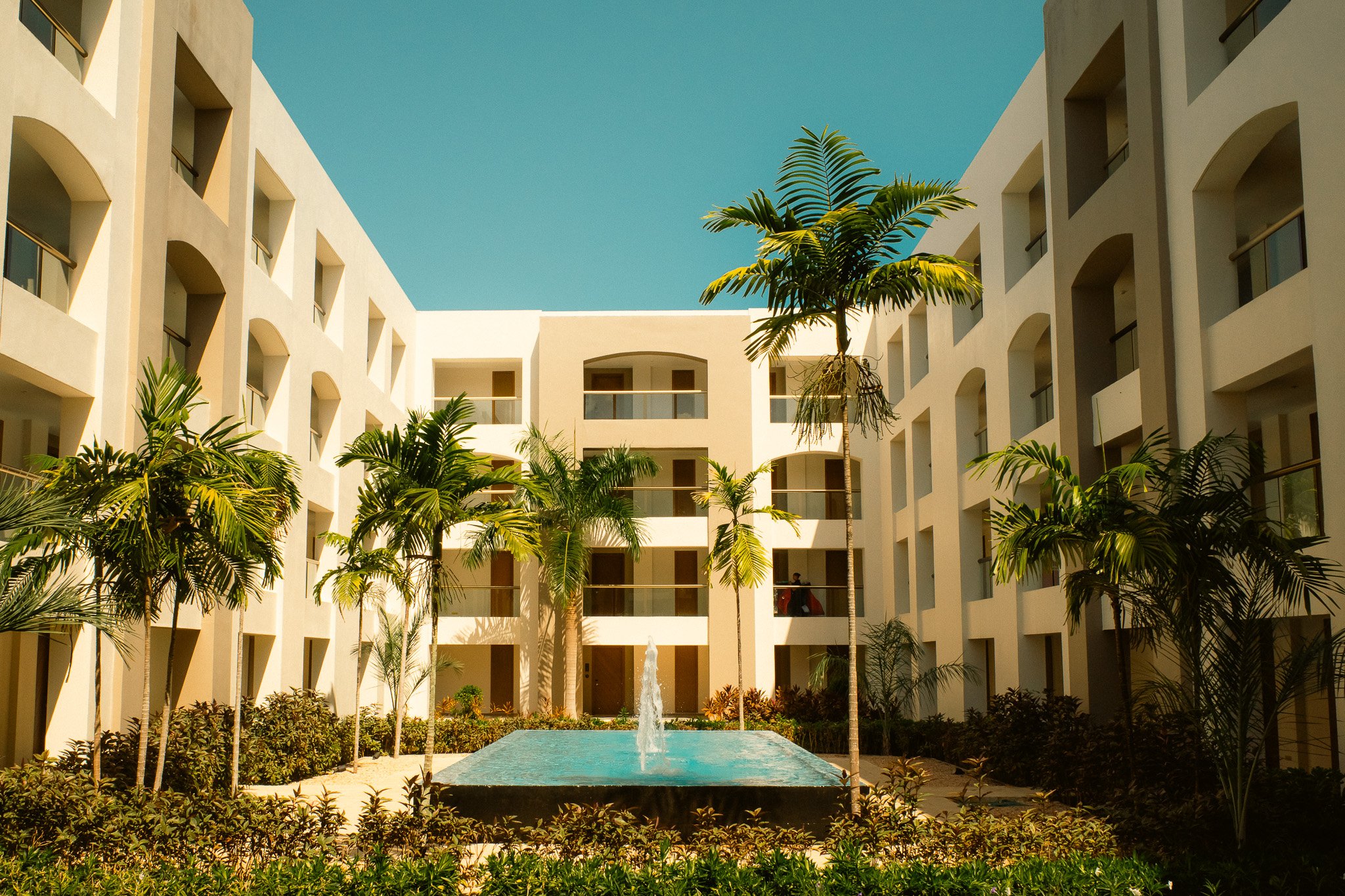 A courtyard with a fountain surrounded by tropical palm trees, situated between white multi-story residential buildings with balconies, under a clear blue sky.