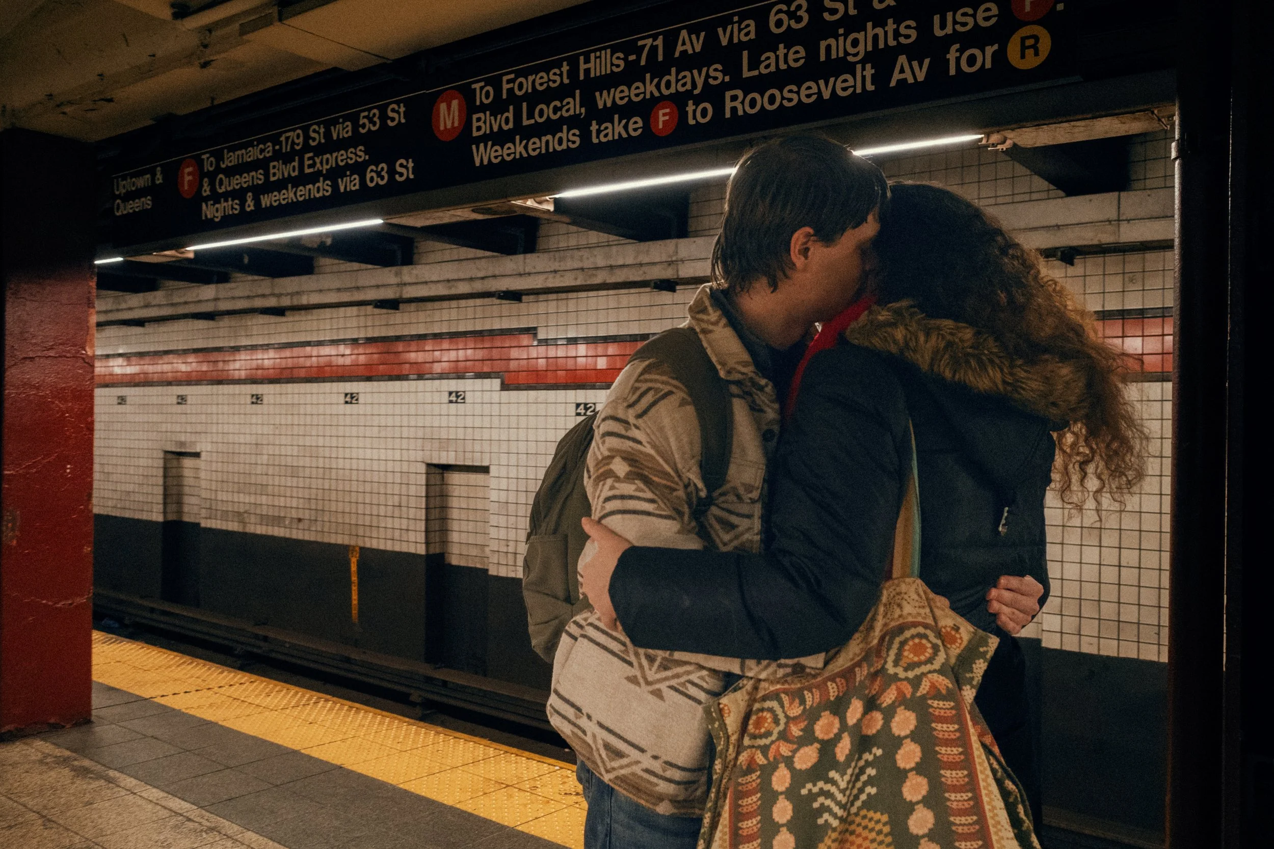 A couple sharing a kiss and hugging on a subway platform, with a subway station wall and sign in the background.