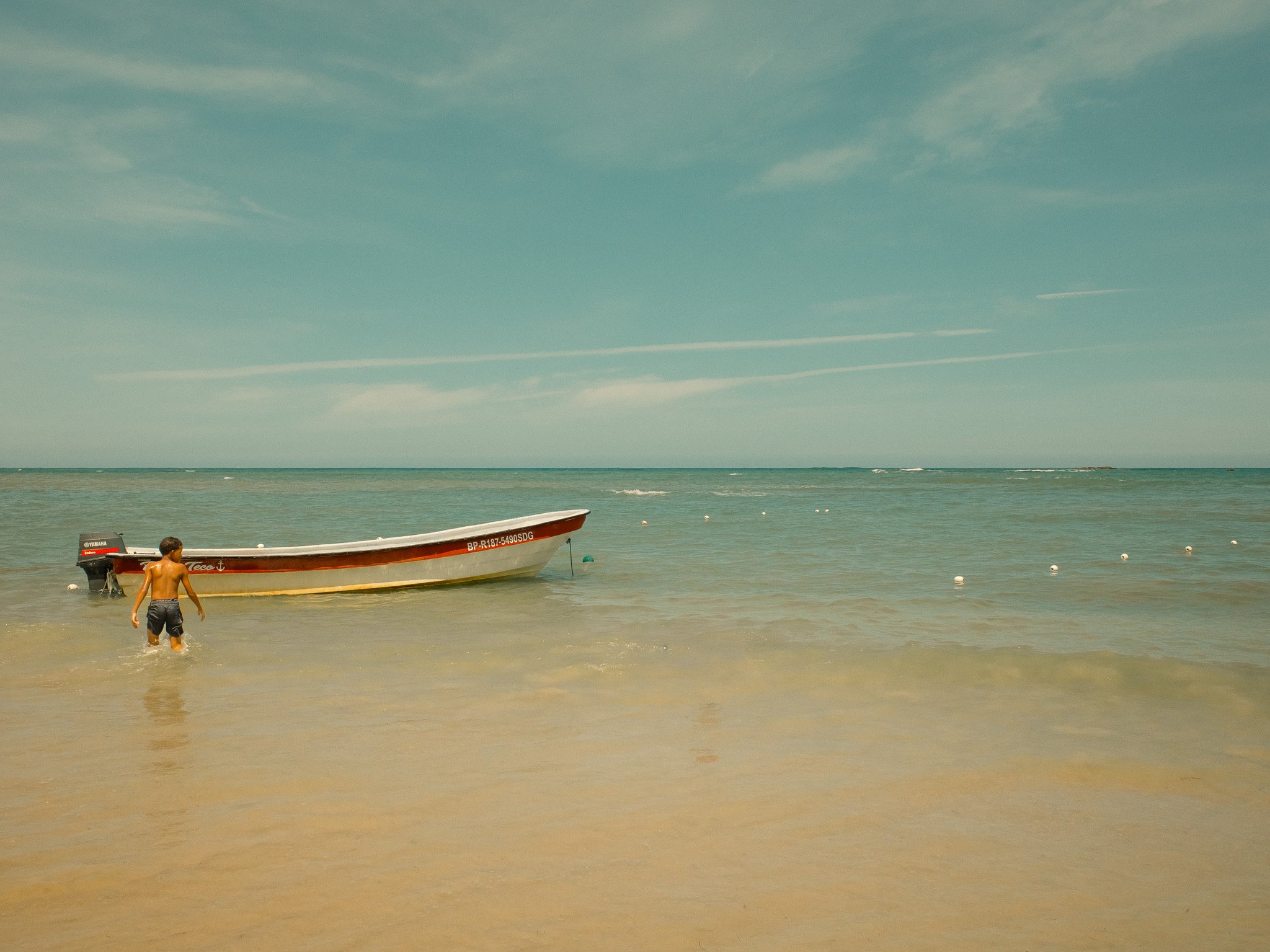 Child wading in shallow beach water near a small boat with outboard motor, facing toward the boat, with several buoys floating in the distance under partly cloudy sky.