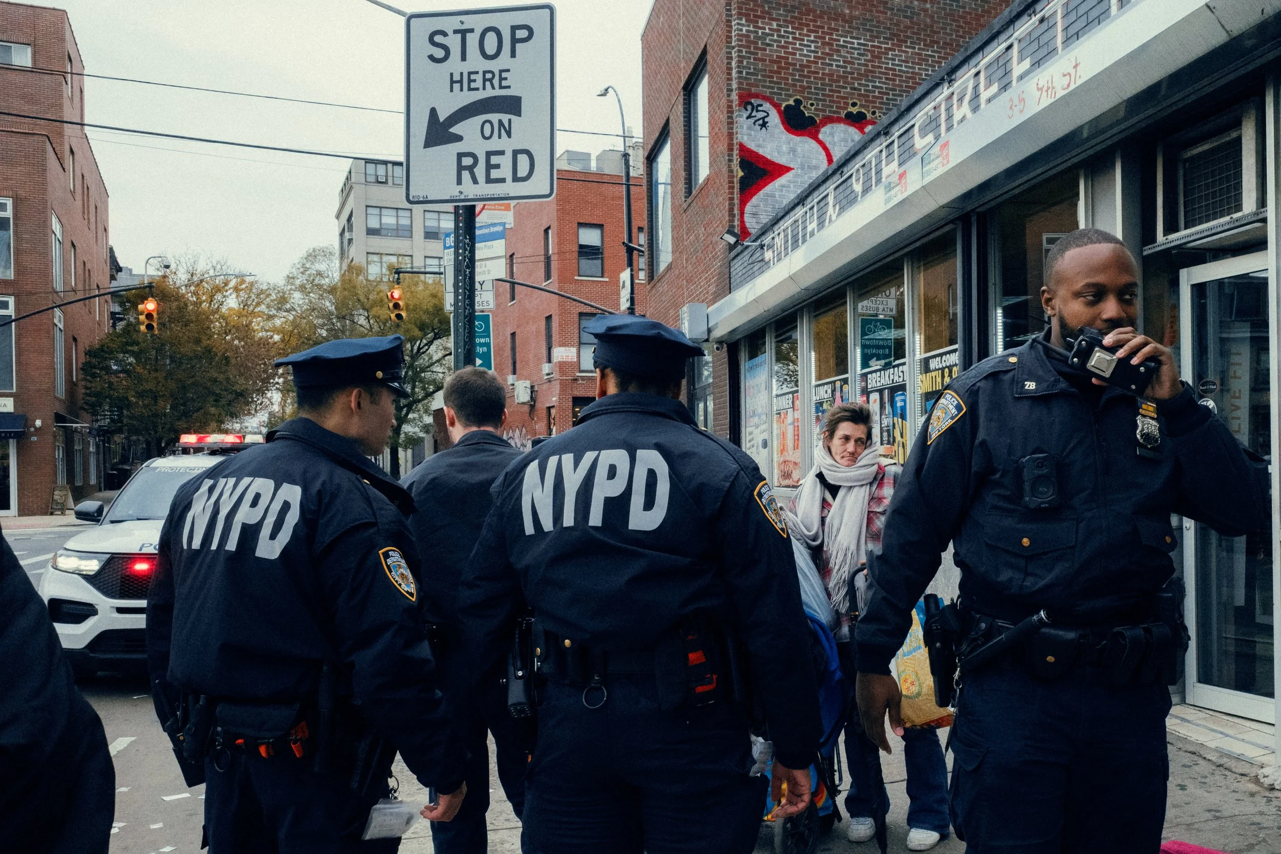 NYC police officers gathering on a city street near a store, with one officer using a radio, and a woman walking nearby, in an urban area with brick buildings and a stop sign.