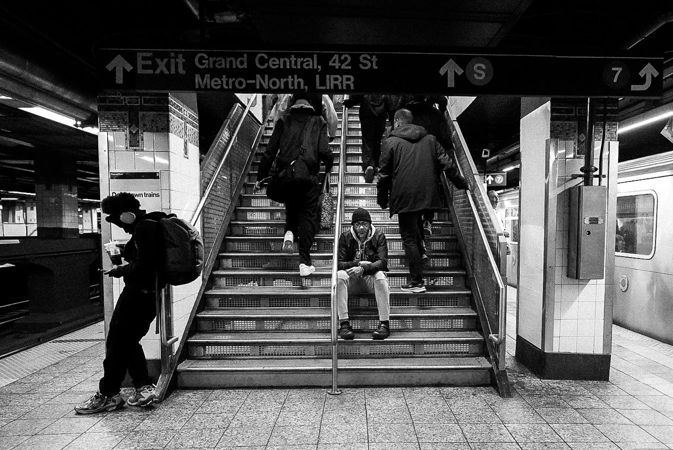 People on stairs at a subway station, with a sign overhead indicating the exit to Grand Central, 42nd Street, and the Metro-North, LIRR trains, and directions to trains 7 and S.