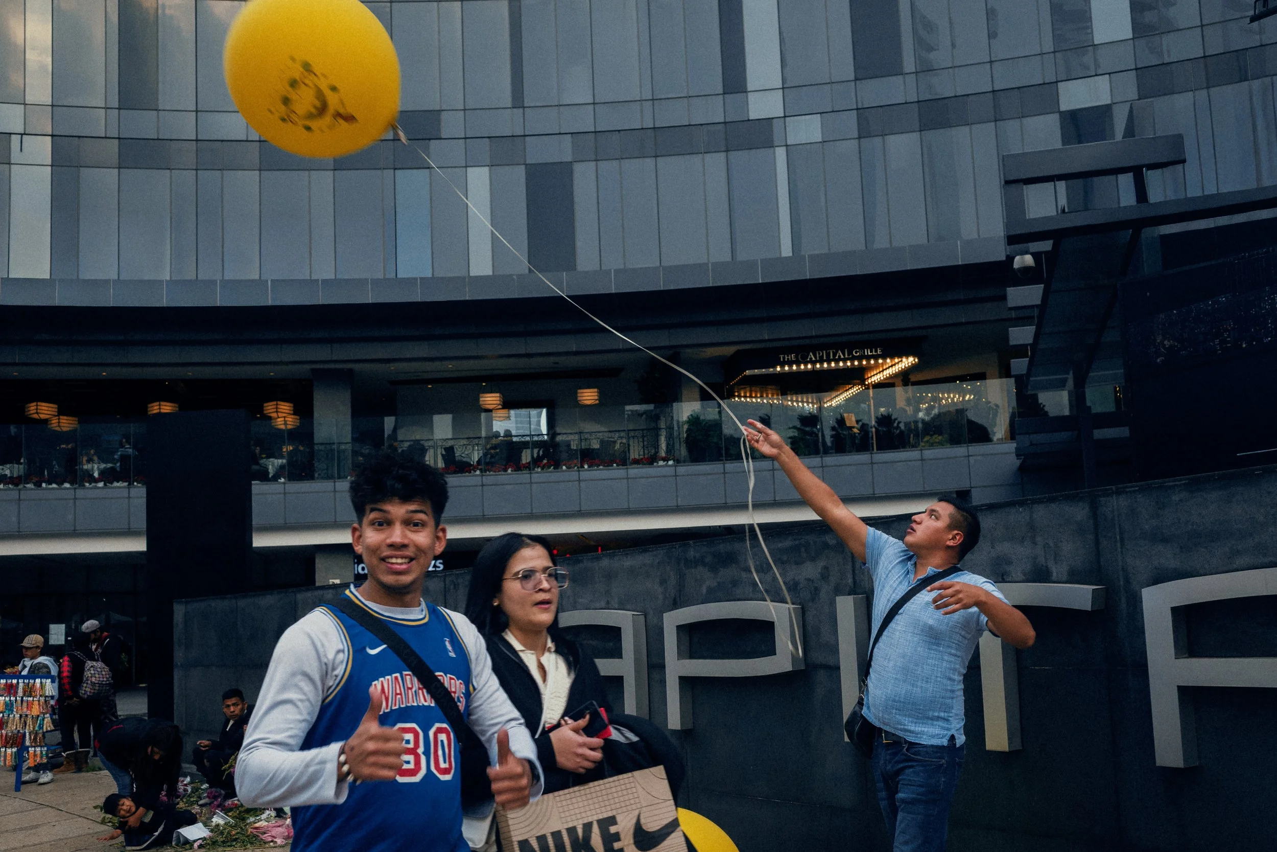 Three people standing outside a modern building with large glass windows. One young man in a blue sports jersey is giving a thumbs-up, a woman with glasses holding a shopping bag, and another man is reaching up to throw a yellow balloon into the air.
