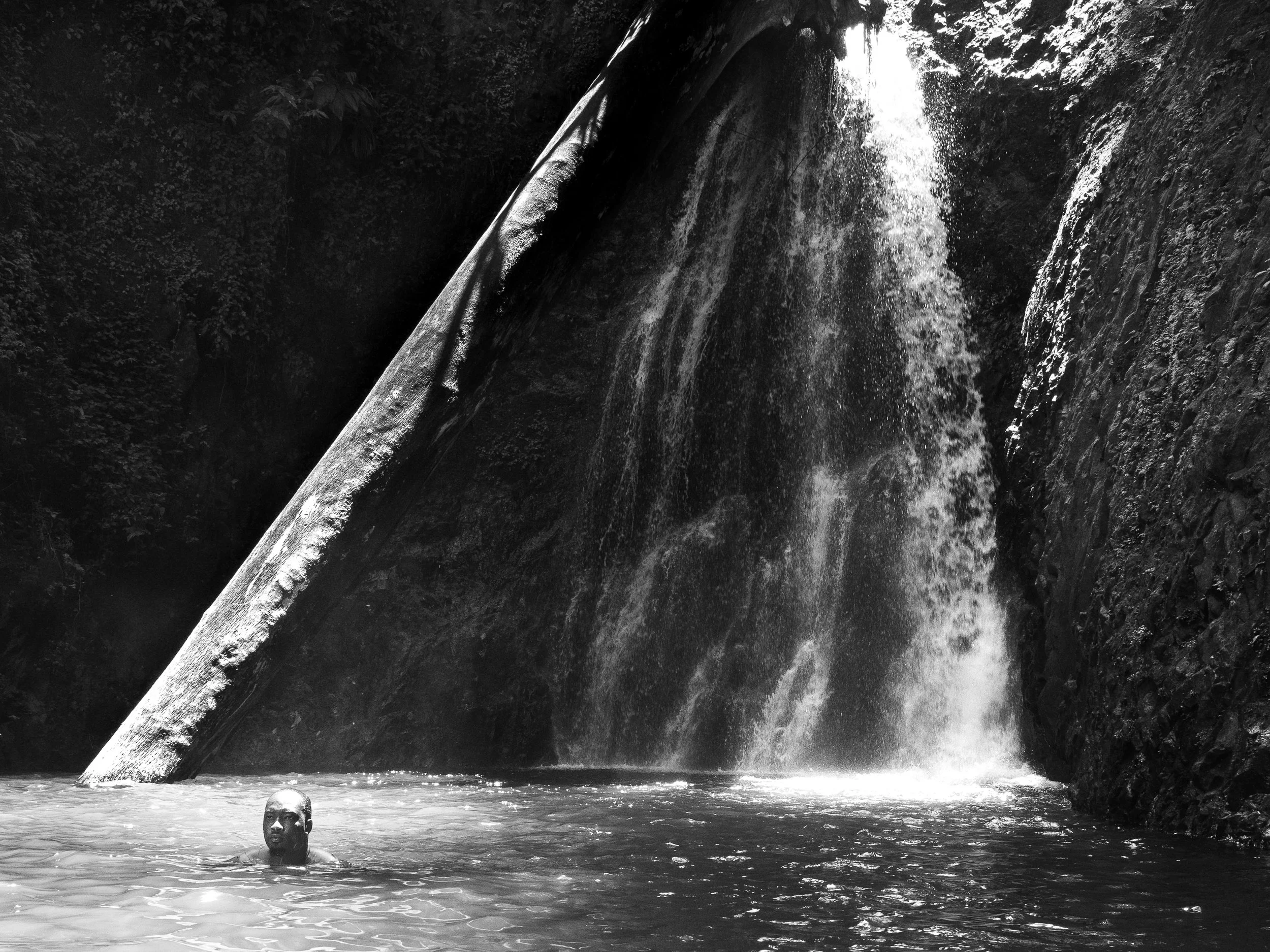 A man swimming in a pool of water beneath a waterfall with a large rock overhang and lush vegetation.