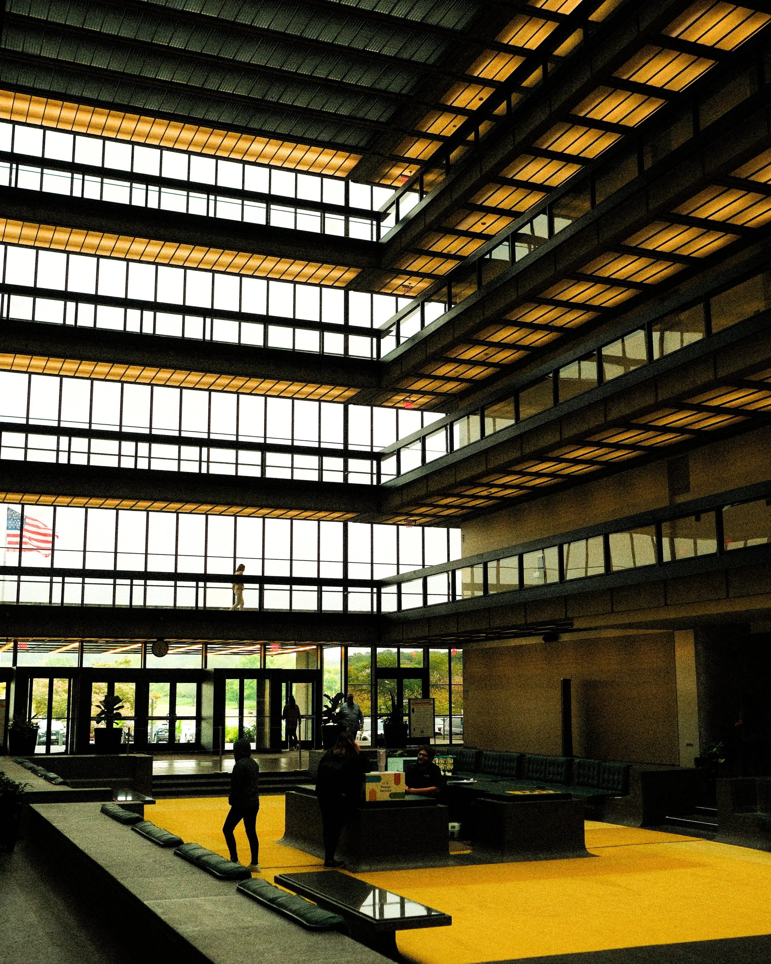 Interior of a modern building with large glass windows, a high ceiling, multiple floors, and a yellow carpeted area where people are walking and talking.