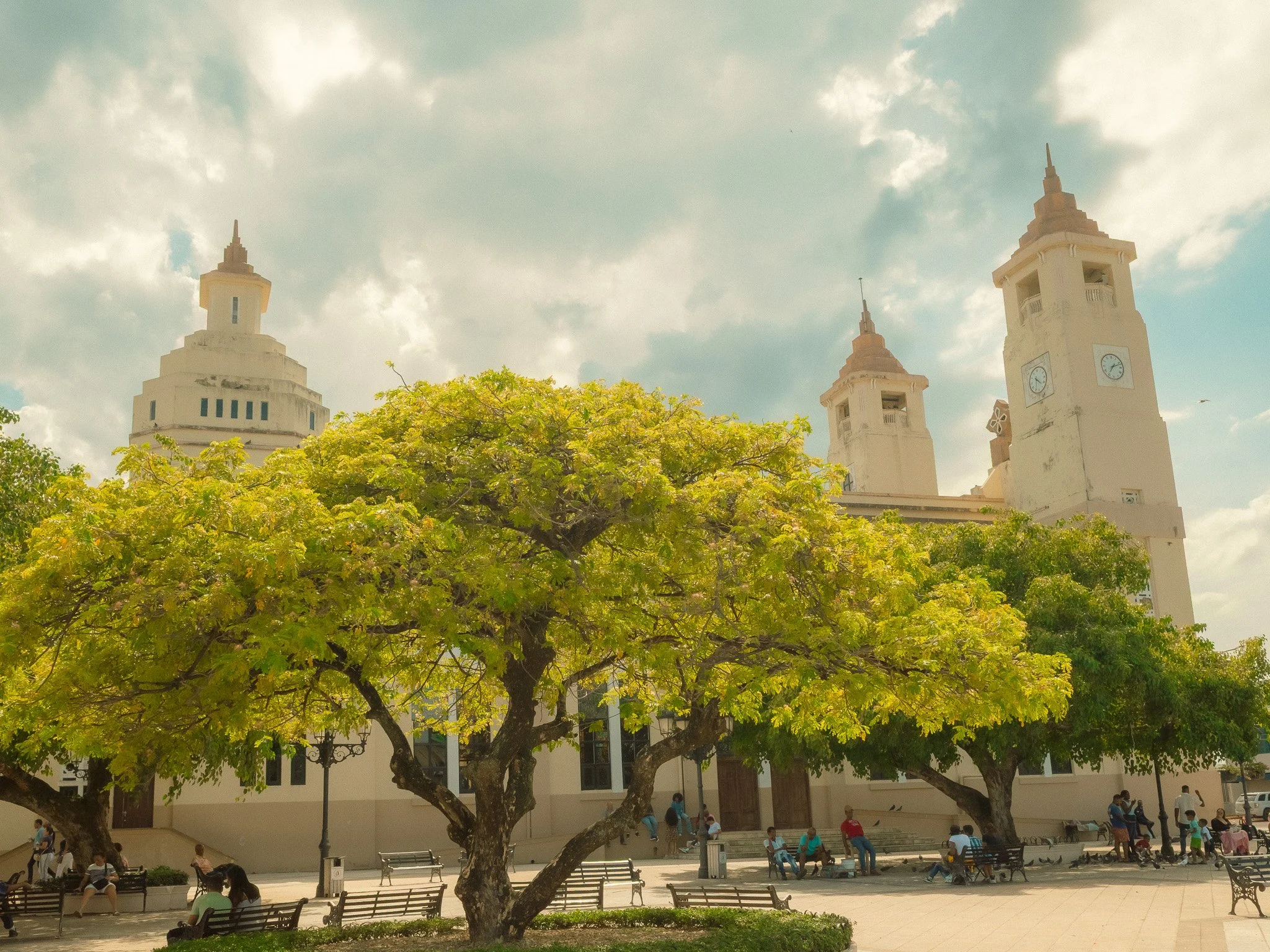 A city square with a large leafy green tree in the foreground, benches, and people sitting and standing. In the background, there is a large white building with three clock towers and a cloudy sky overhead.
