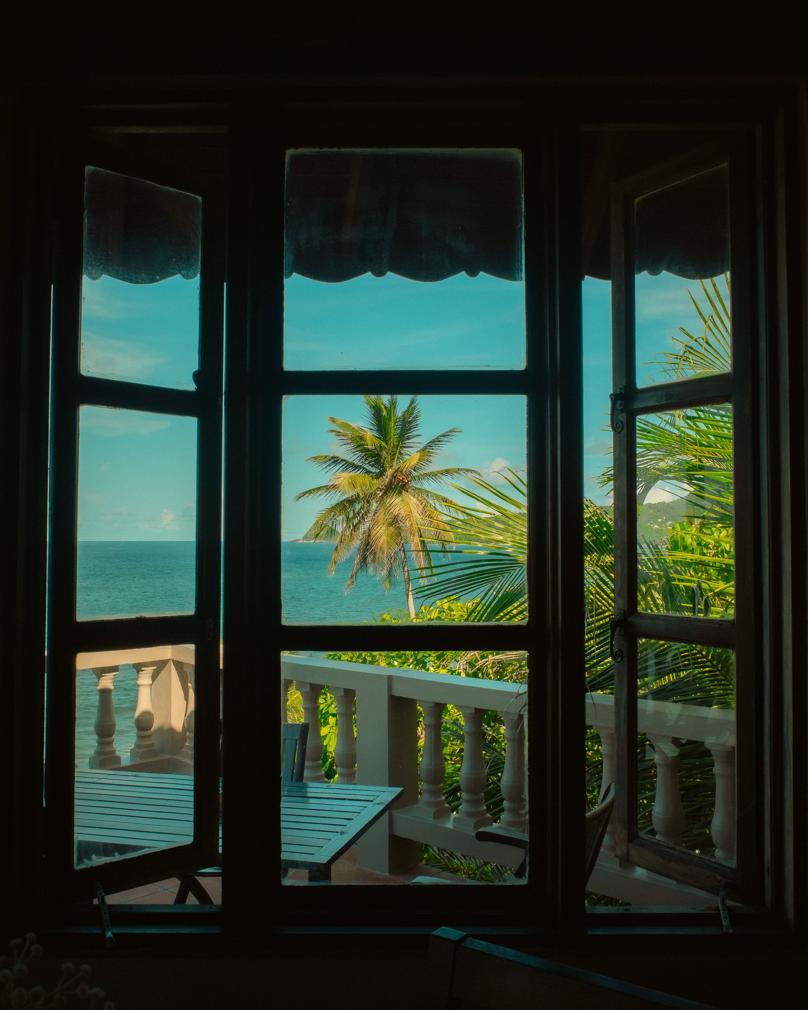 View of a beach scene through open window with palm trees, ocean, and balcony with white railing