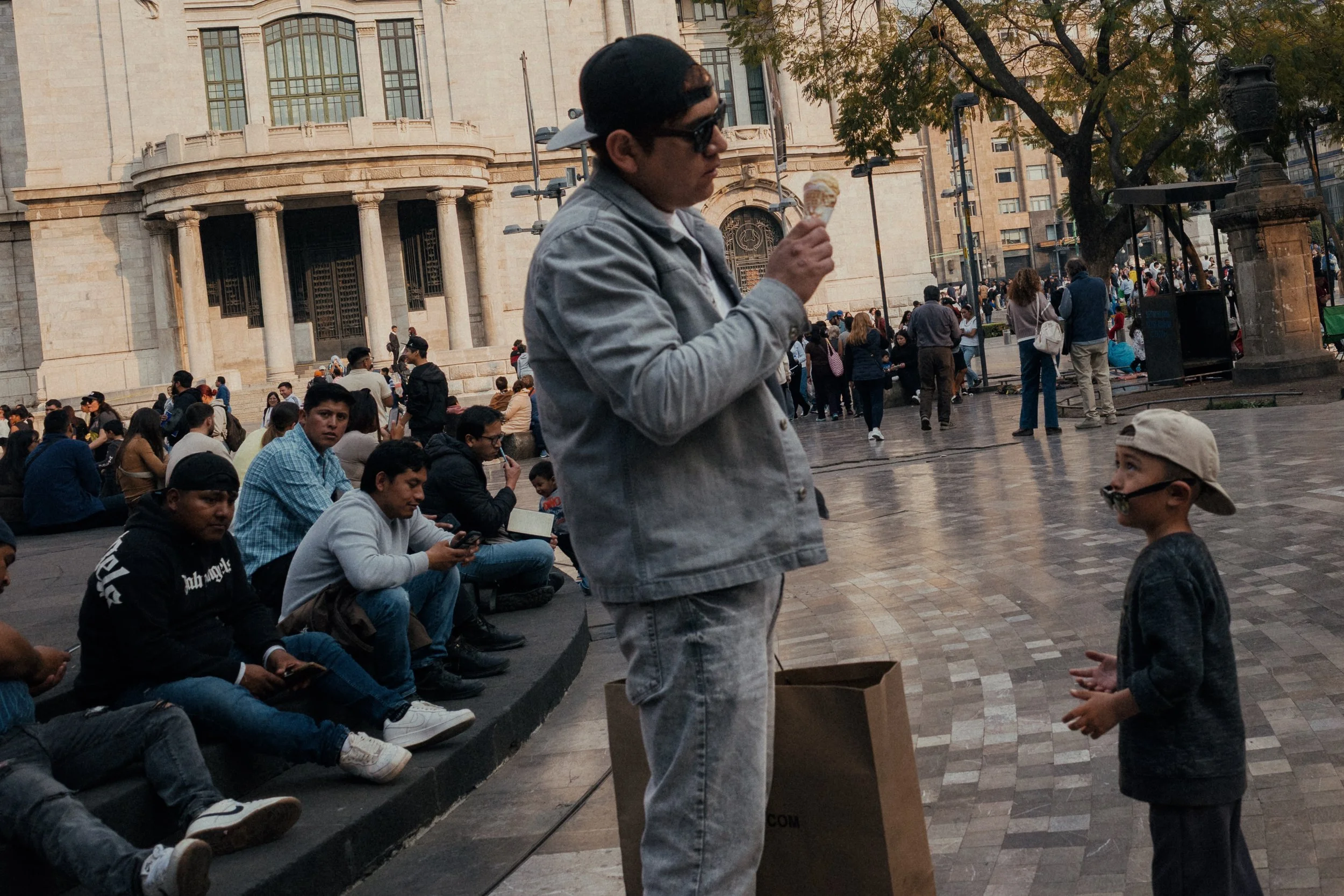 A man in sunglasses and a cap standing with a paper bag, eating ice cream, talking to a young boy wearing a cap and dark glasses, at a busy outdoor plaza with many people, trees, and historic buildings in the background.
