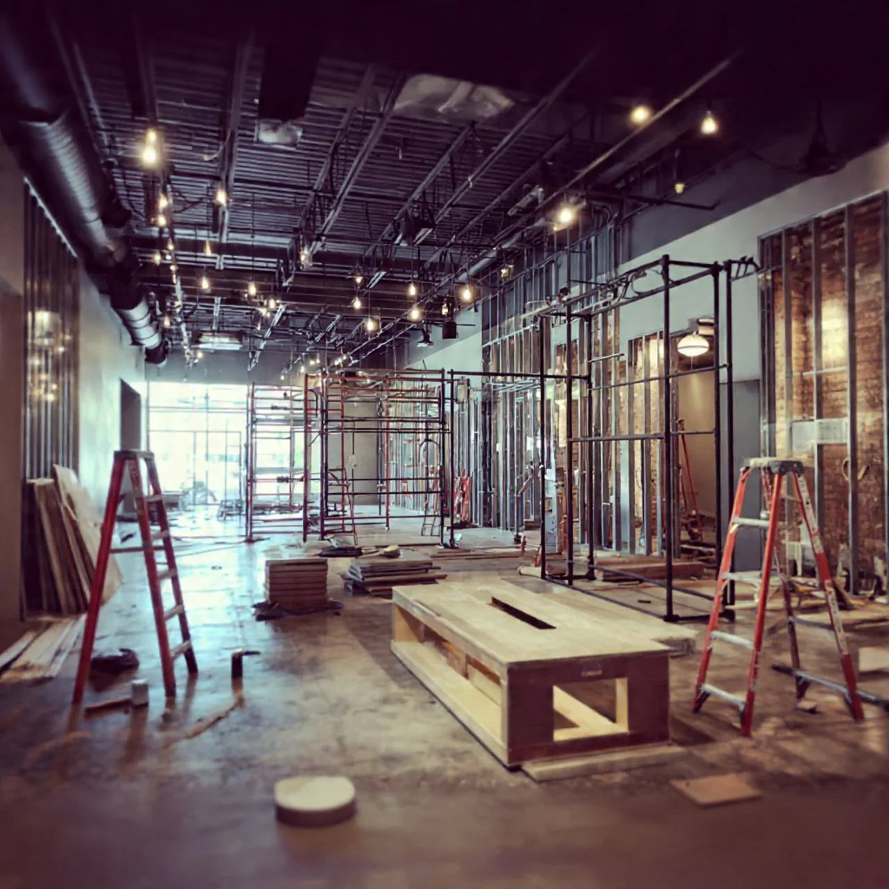 Interior of a building under construction with scaffolding, ladders, construction materials, and exposed ceiling pipes and wires.