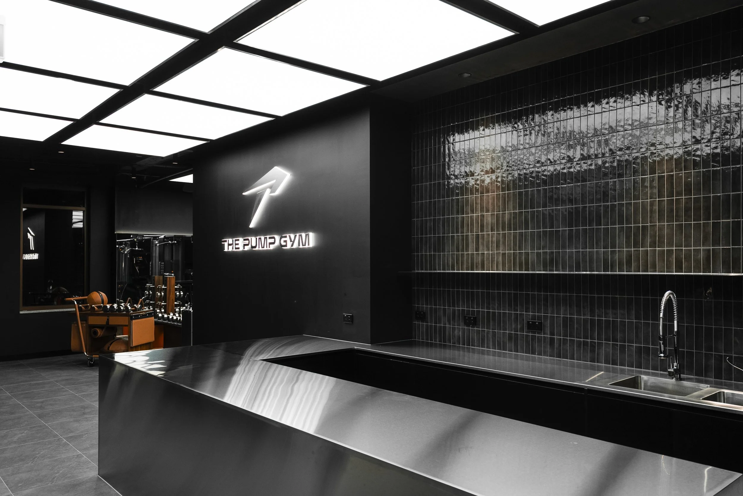 Modern fitness studio with black walls, illuminated sign reading 'The Pump Gym', black tiled wall, and sleek metal countertop. Exercise equipment visible in the background.