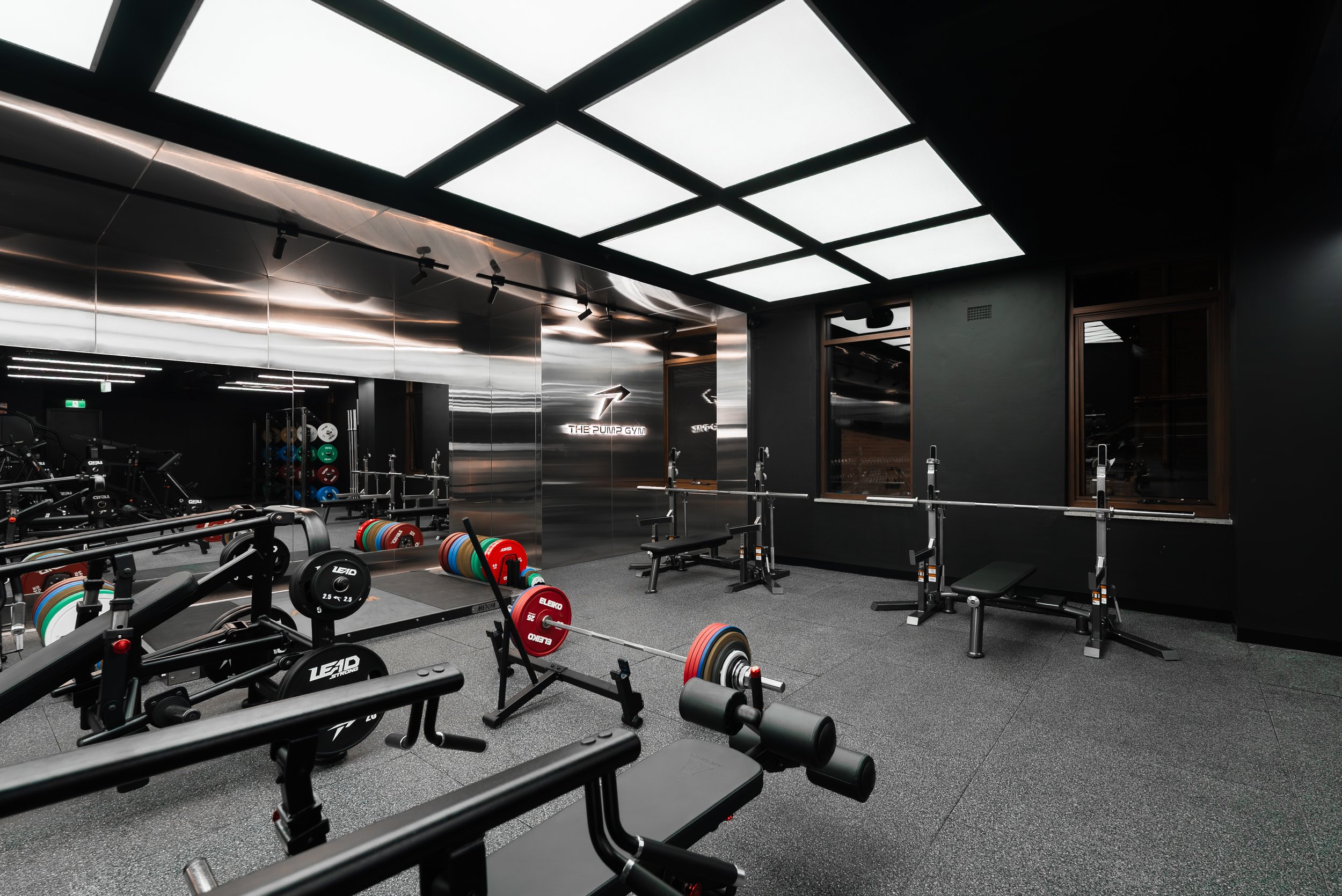 Modern gym with black walls, large illuminated ceiling panels, weightlifting equipment, and a mirror wall with a sign that reads 'The Pump Gym'.