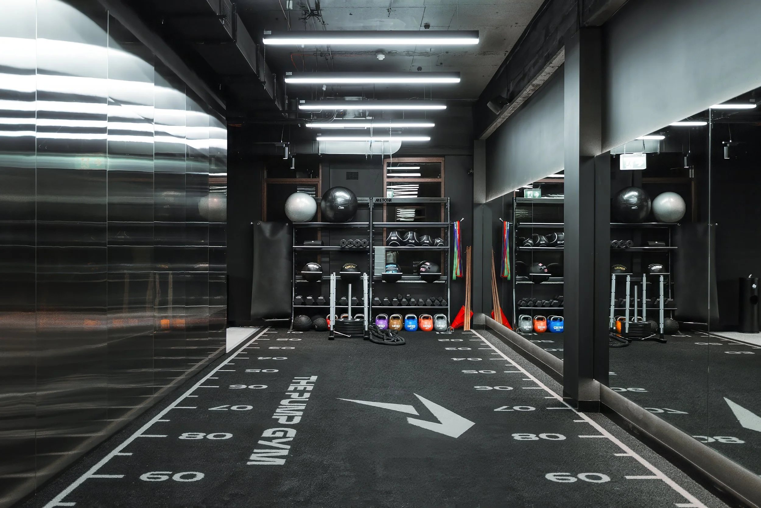 A fitness gym with black walls, a black carpet with white markings for exercises, and various workout equipment such as medicine balls, kettlebells, resistance bands, and ropes organized on shelves. The ceiling has exposed pipes and long fluorescent lights.
