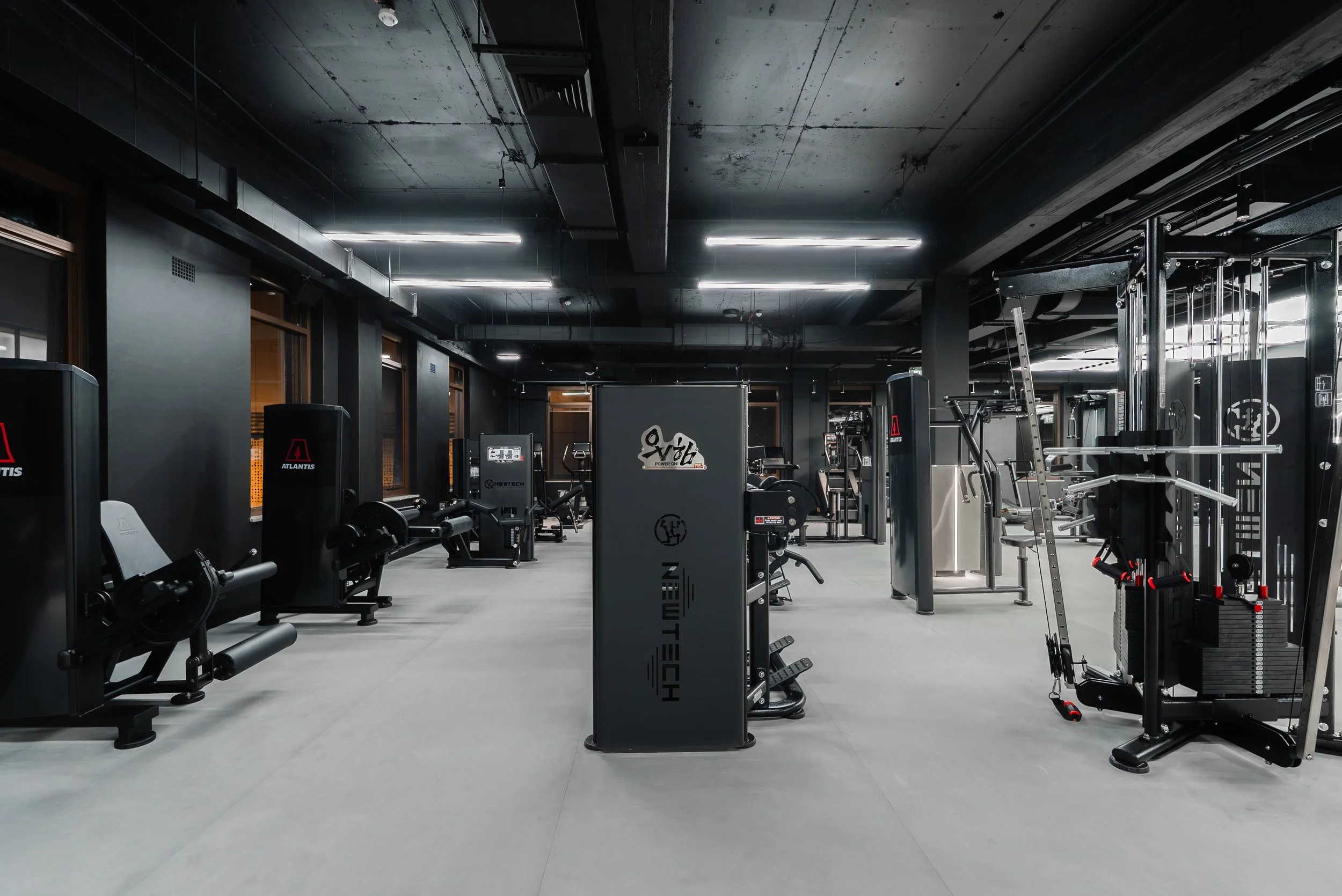 Interior of a modern gym with various workout machines and equipment, dim lighting, black ceiling with exposed pipes and ducts, and large windows along the wall.