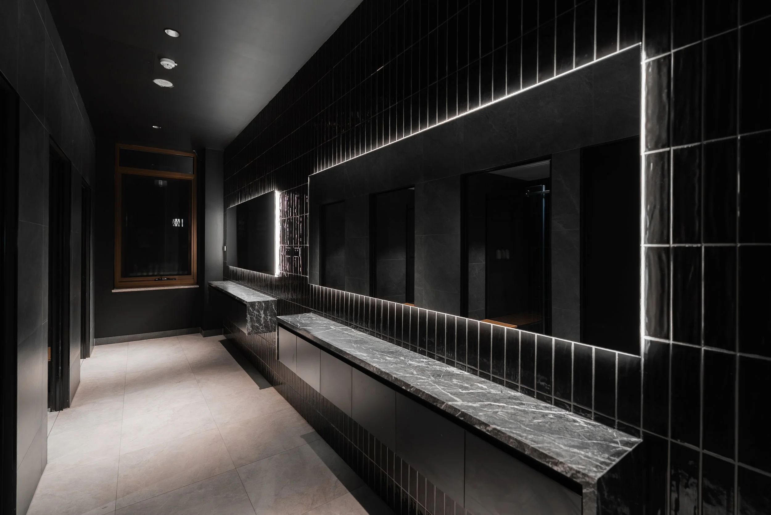 Dark bathroom with black tiled walls, a black countertop with marble top, and a large mirror with backlit LED lighting.