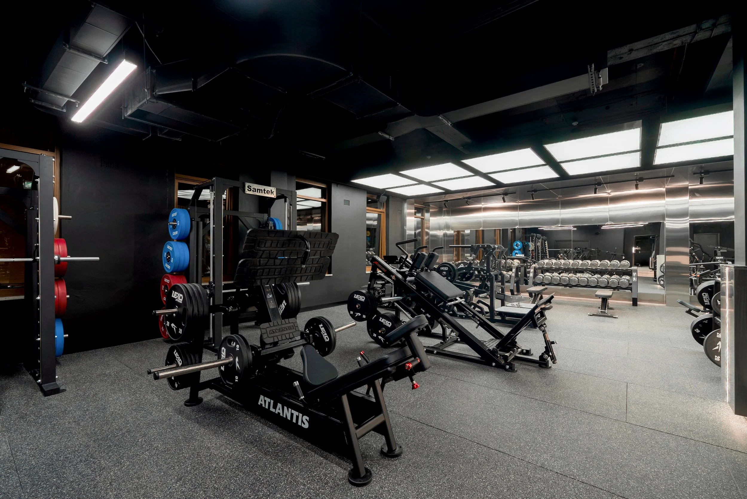 A modern gym with various workout machines, free weights, and lifting equipment. The lighting is bright, and the space has a sleek, minimalistic design with dark and reflective surfaces.