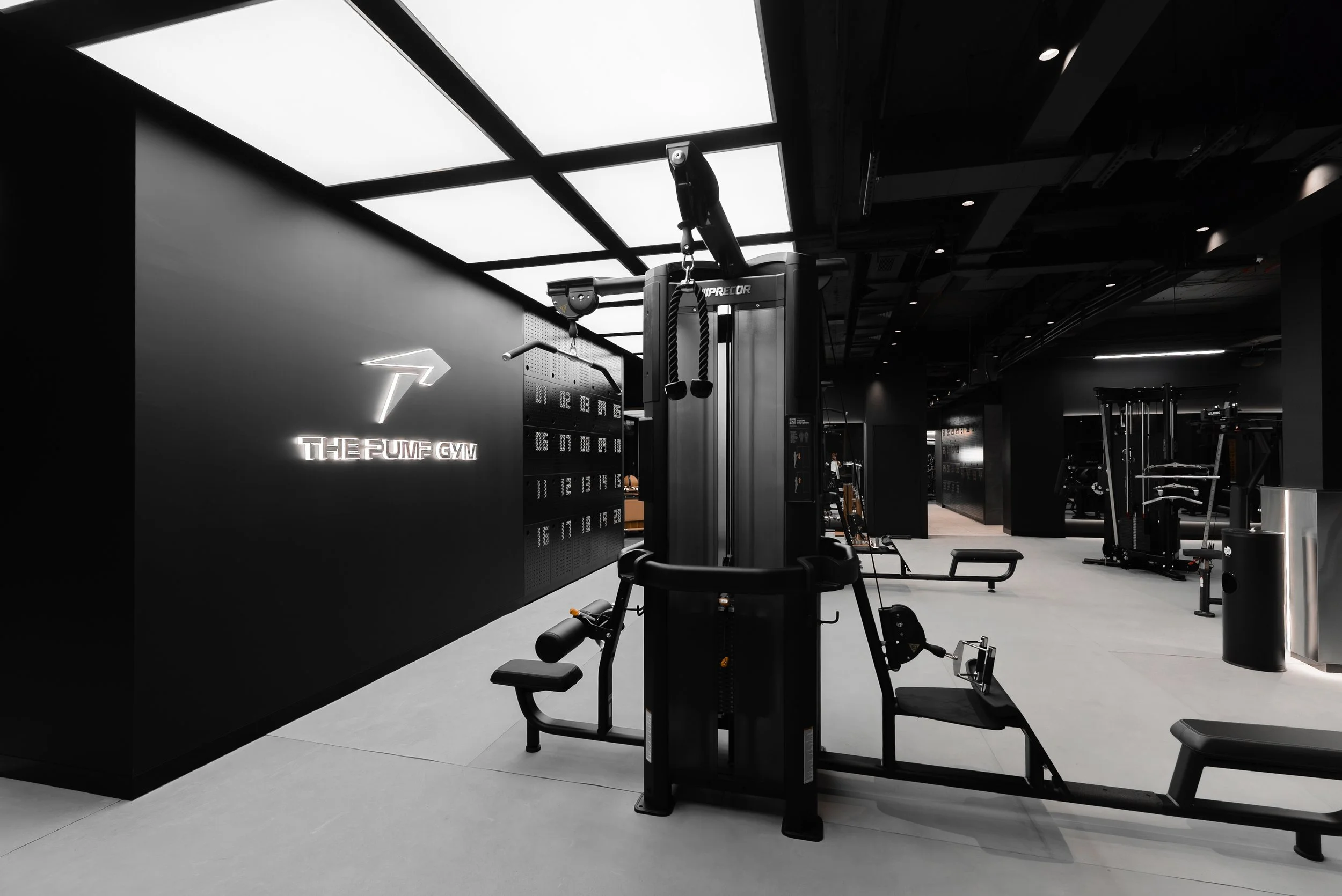 A modern gym with exercise equipment, including a multi-function weight machine, benches, and other fitness machines. The space has a sleek, black and gray interior with ceiling lights and a sign that reads 'THE PUMP GYM' with a lightning bolt logo.