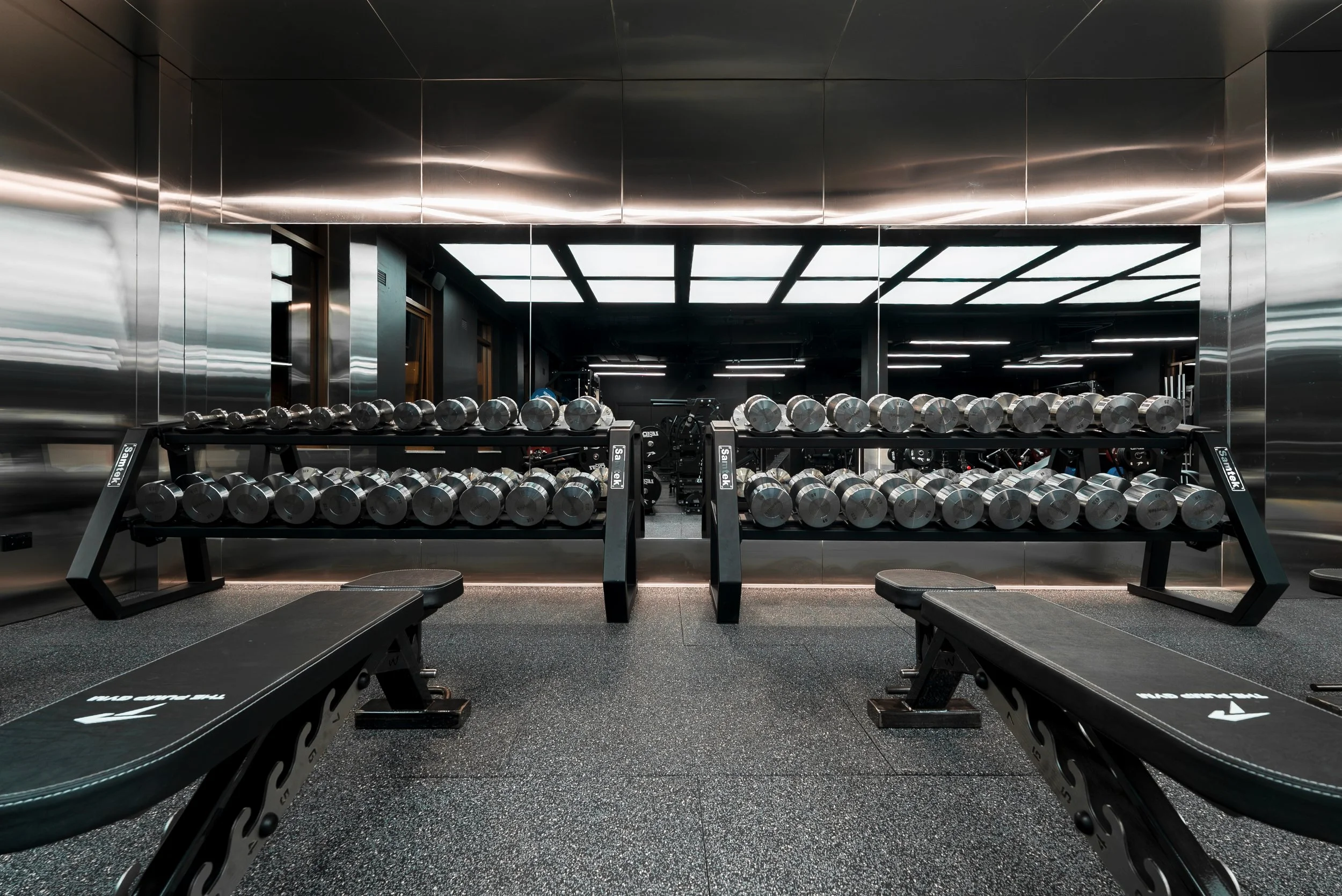 Gym with black rubber flooring, two benches, and a wall of dumbbells reflected in a large mirror. The ceiling has bright rectangular lighting panels.