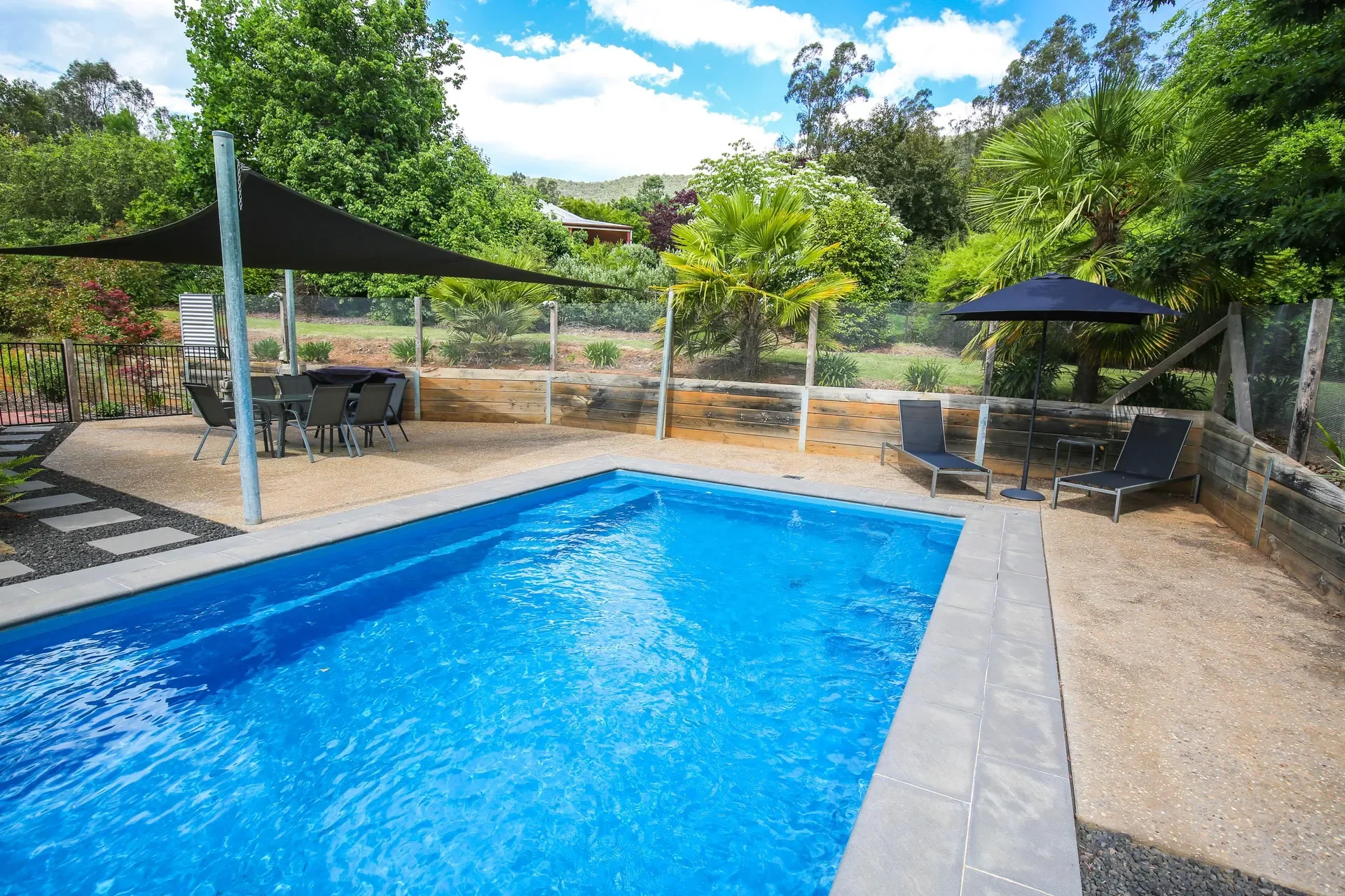 Outdoor swimming pool with blue water, surrounded by a concrete deck, black lounge chairs with umbrellas, and lush green trees and plants in the background.