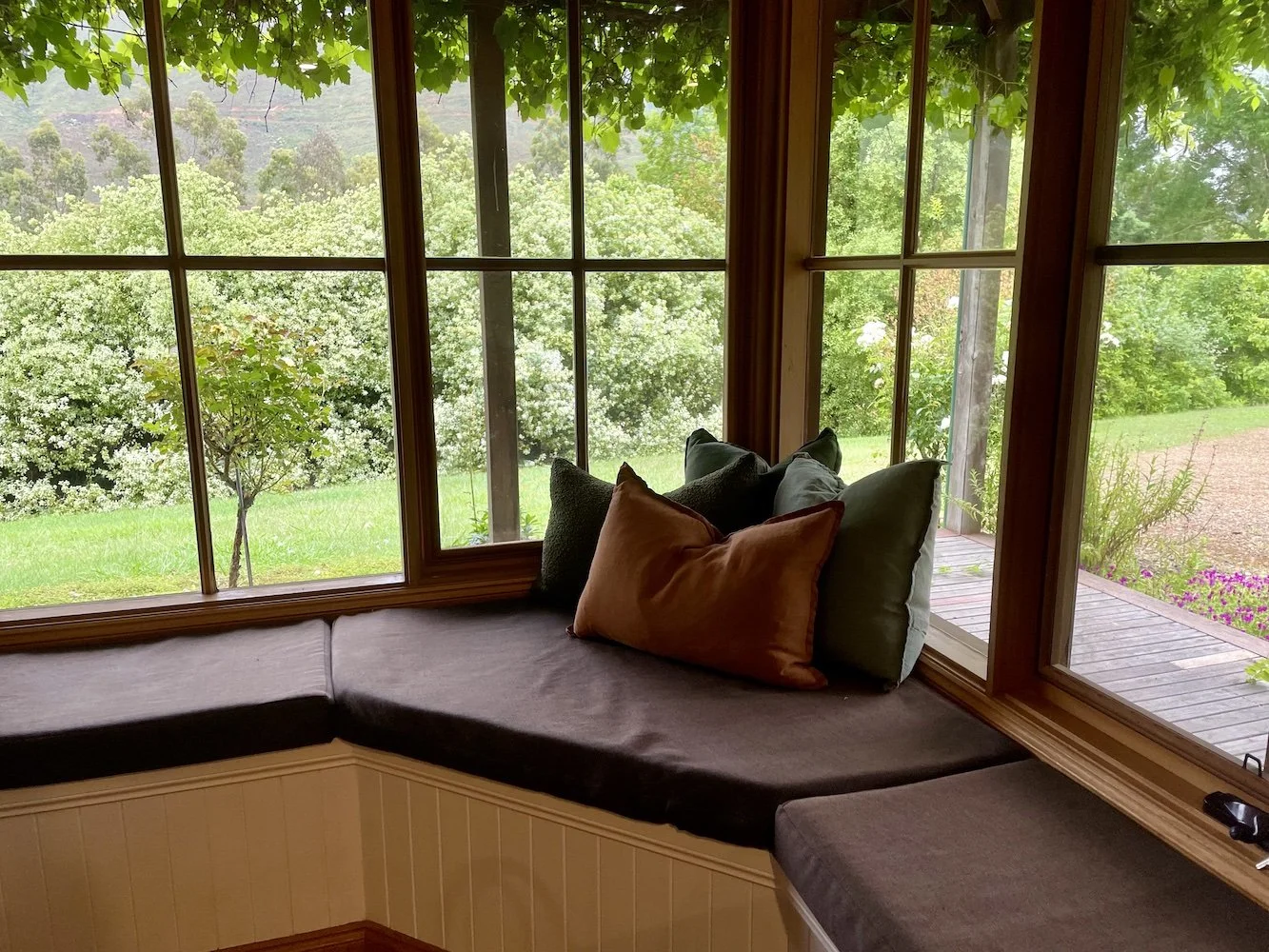 A cozy window seat with cushions overlooking a lush green garden with trees and plants outside.
