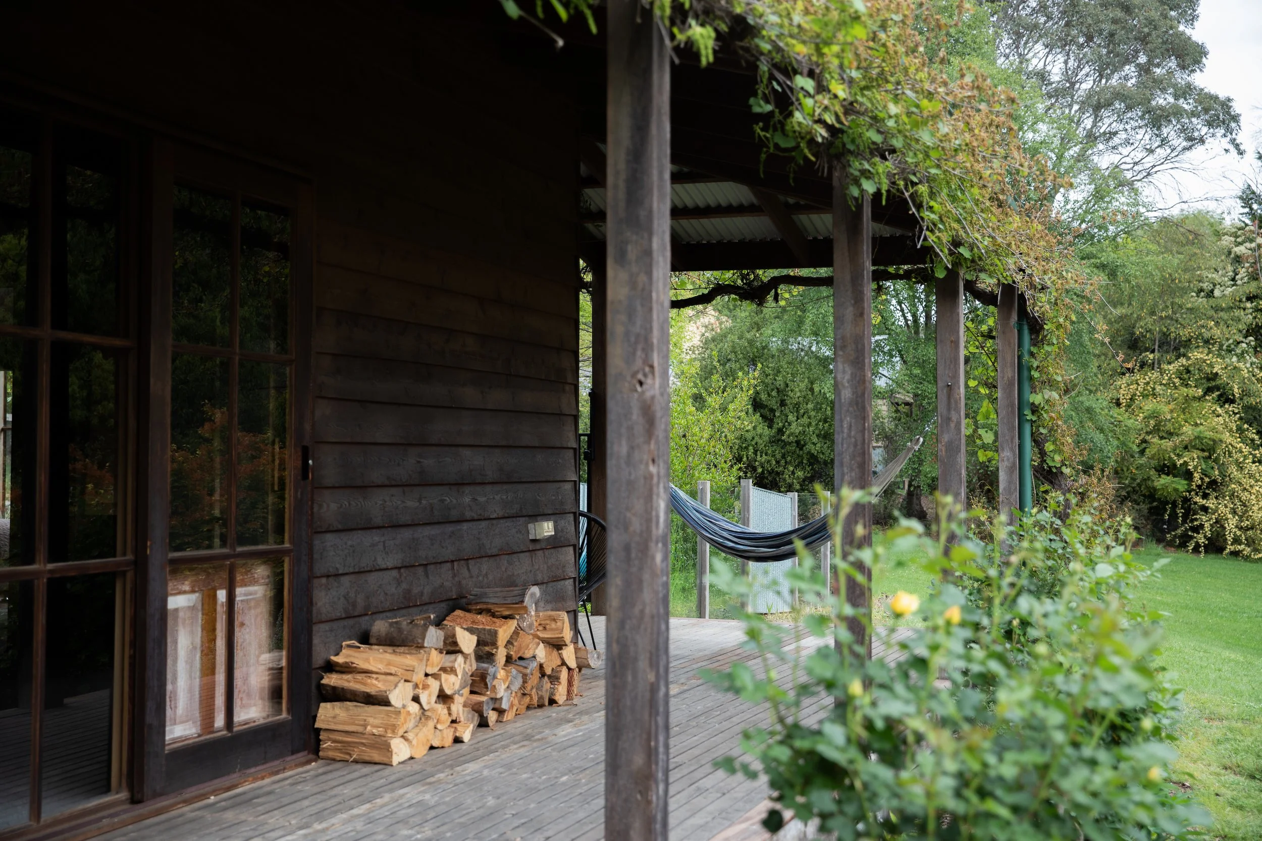 Wooden house patio with stacked firewood, a hammock, greenery, and trees in the background.