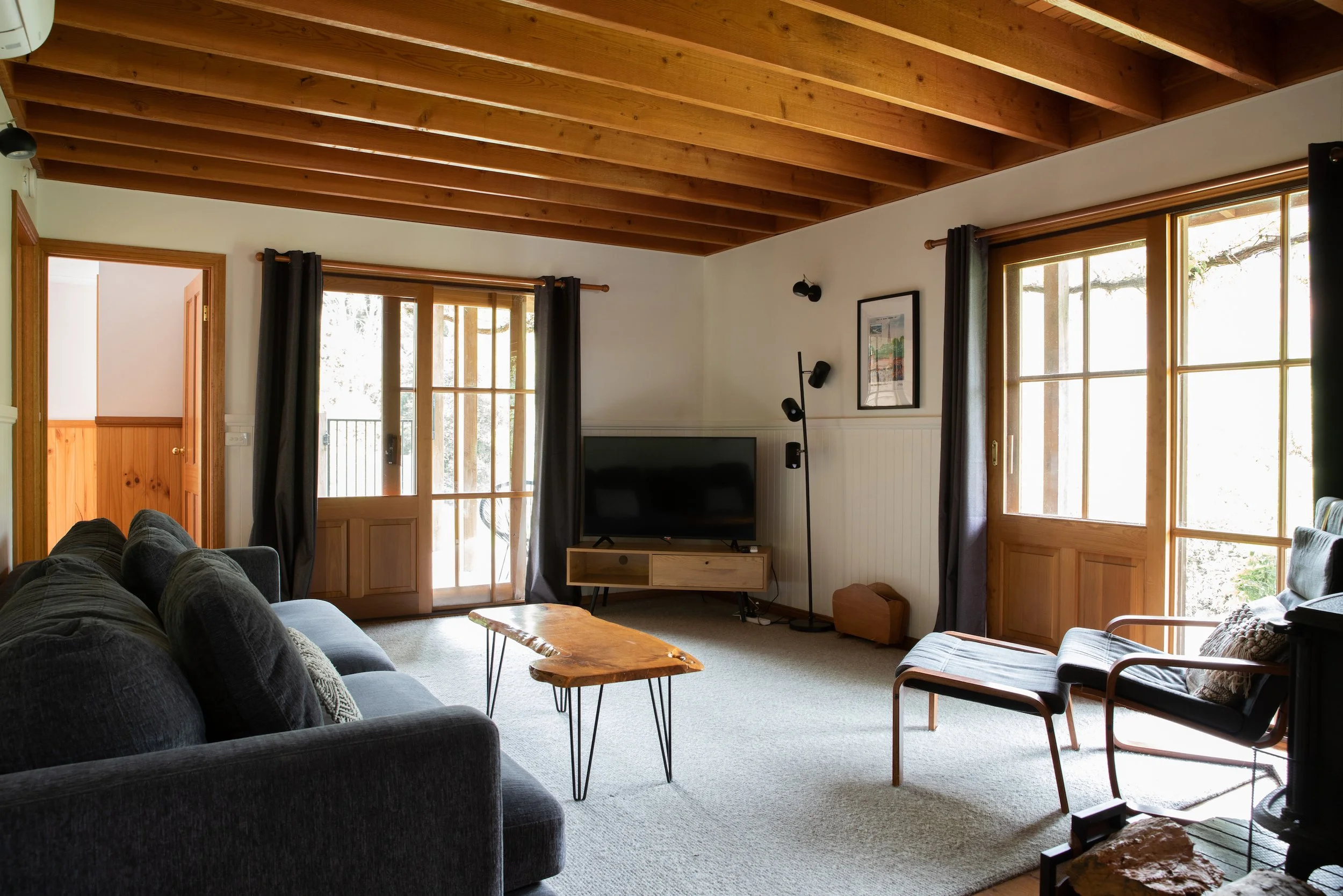 Living room with large windows, wooden ceiling, gray sofa, wooden coffee table, TV on stand, black armchair, and floor lamp.