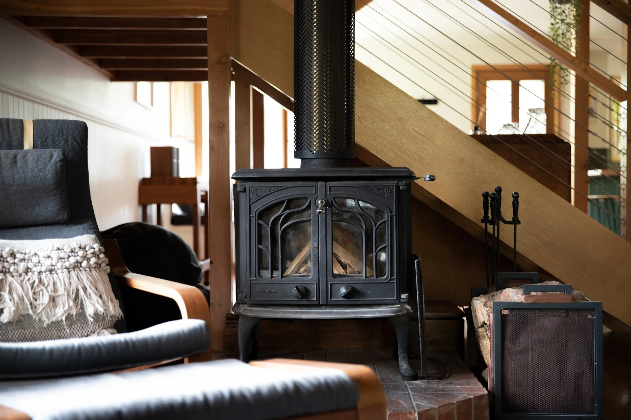 Wood stove, logs, fire tools, a cozy chair with a throw blanket, and a staircase with wooden railings in a home.