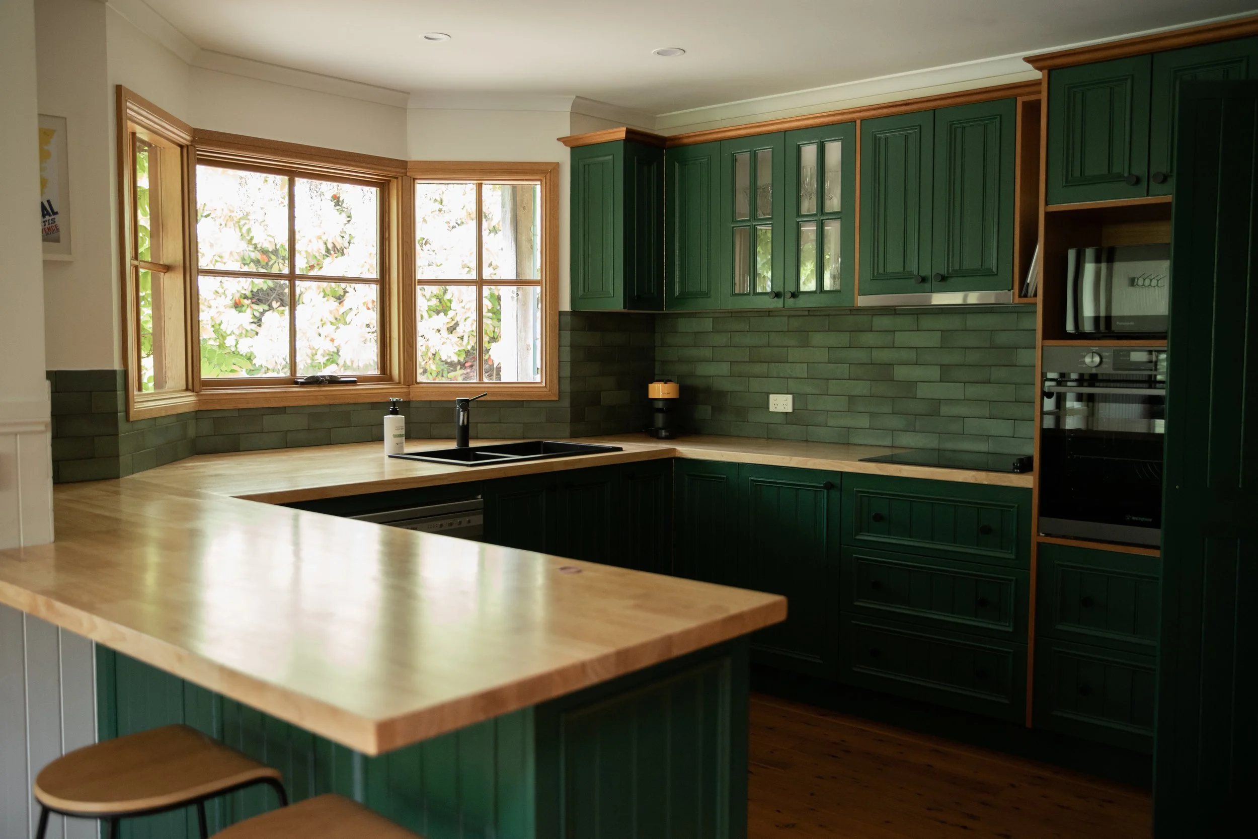 A modern kitchen with green cabinets, light wood countertops, a black sink, and a large window.