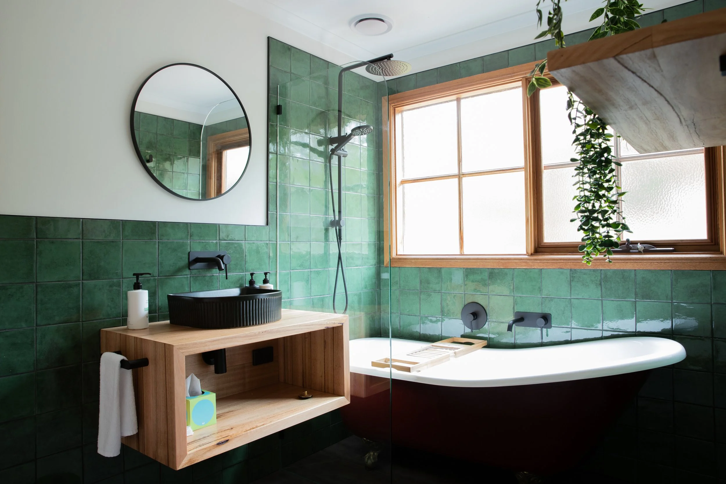 Modern bathroom with green tiled walls, wooden framed window, black sink on a wooden vanity, round mirror, walk-in shower, bathtub, and hanging plants.