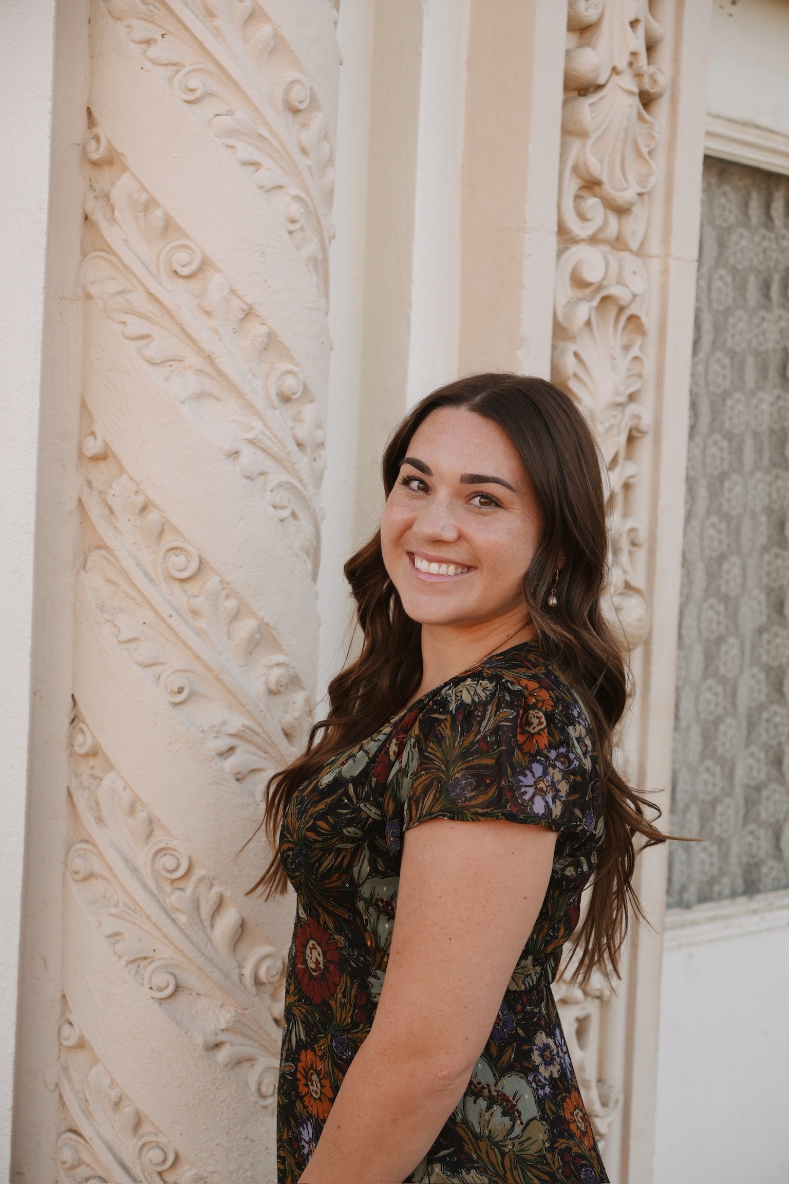 A woman with long brown hair smiling, dressed in a floral patterned dress, standing in front of ornate cream-colored architectural details.