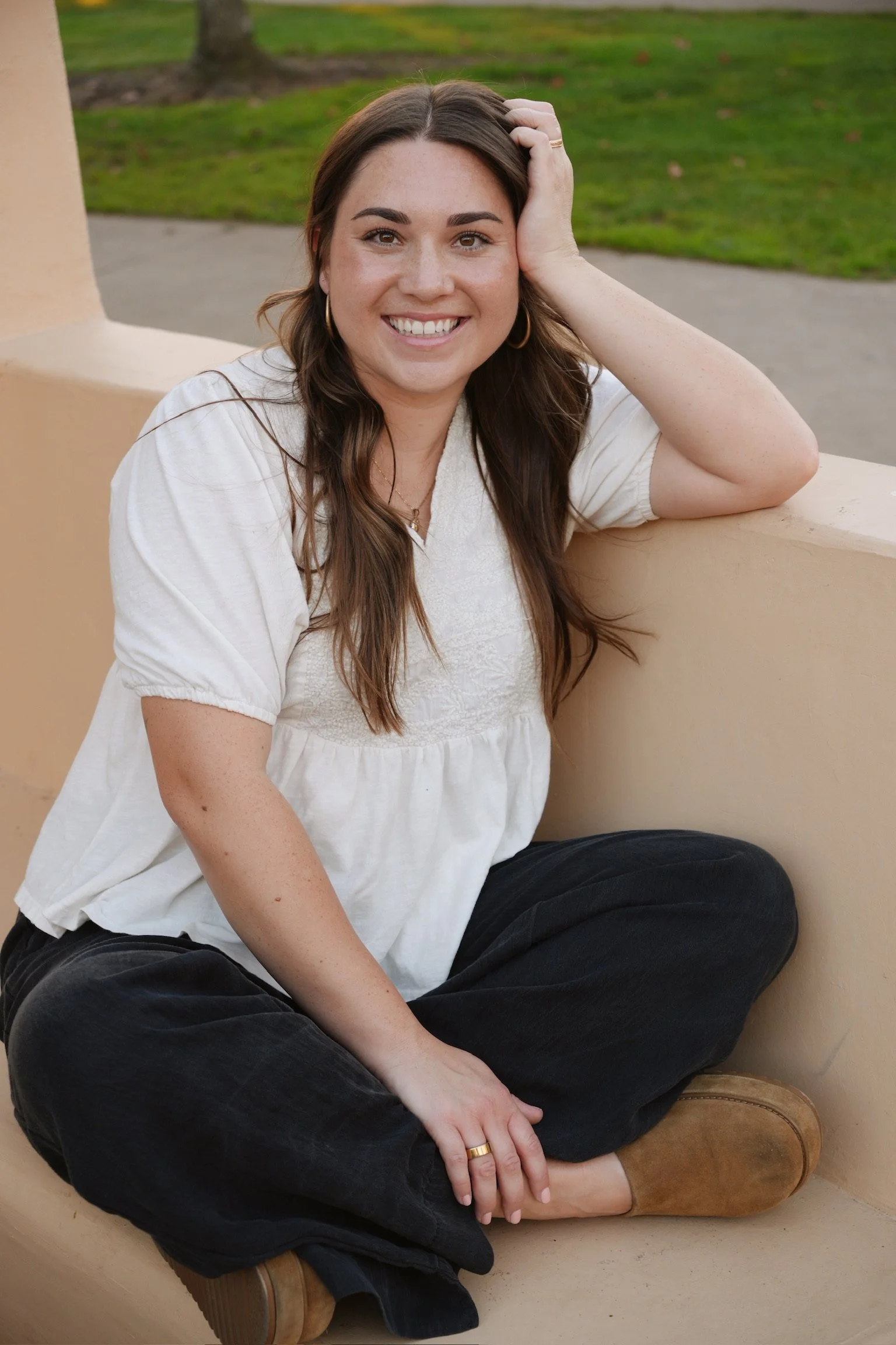 A woman with long brown hair, sitting cross-legged outdoors on a beige bench, smiling at the camera, with a background of grass and trees.