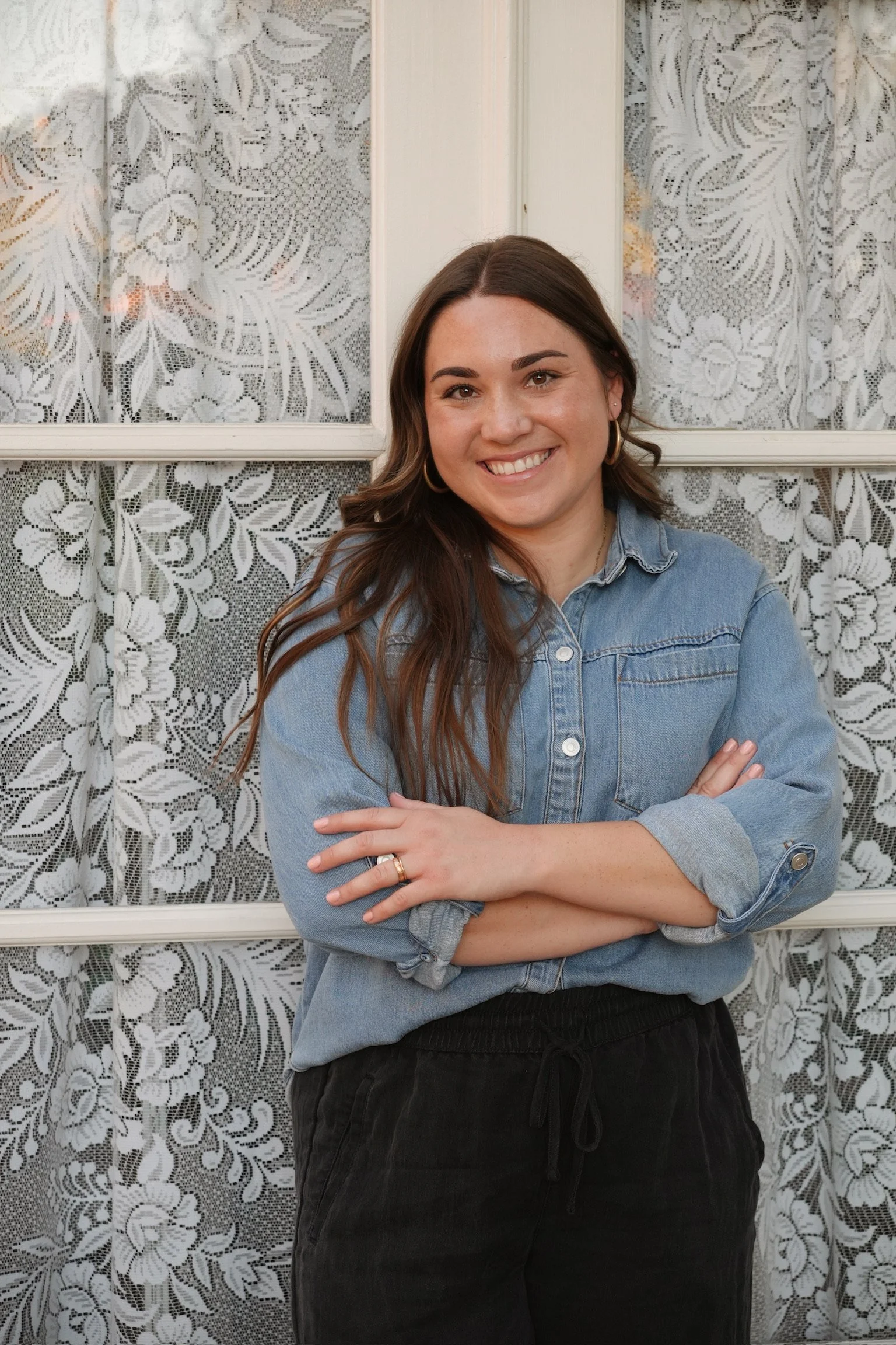 A woman with long brown hair, wearing a denim jacket and black pants, standing in front of lace curtains, smiling with arms crossed.