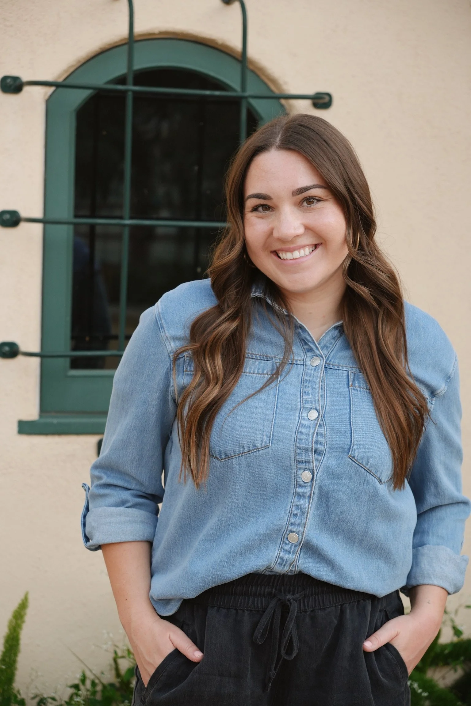 A young woman with long wavy brown hair smiling while standing outdoors in front of a beige wall with a green window and some plants at the bottom.