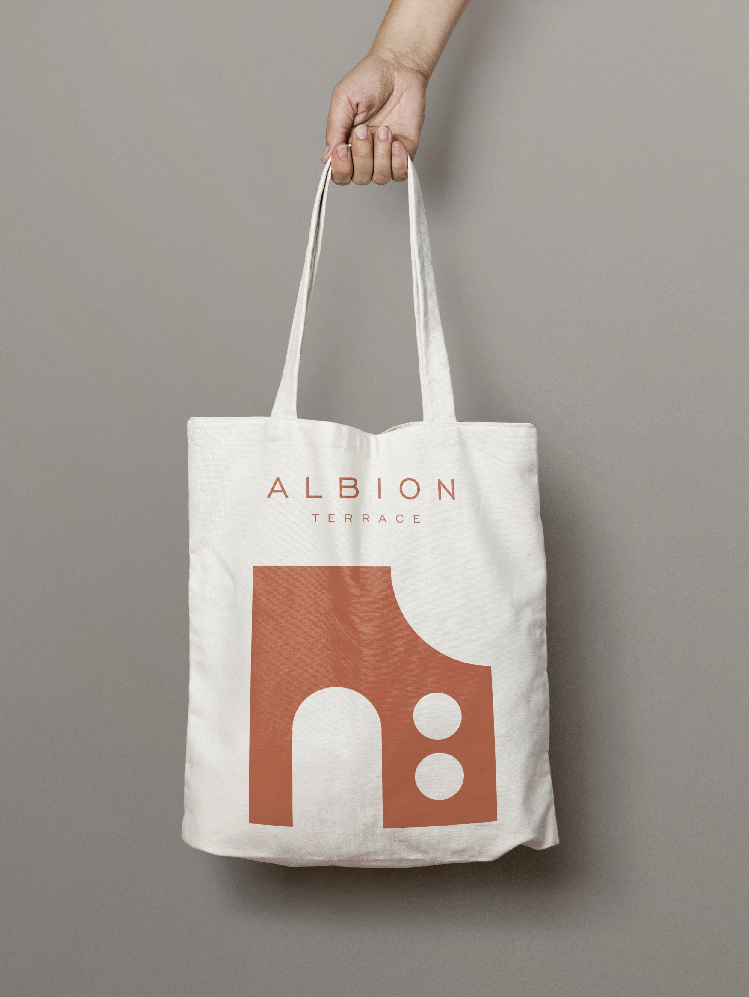Hand holding a white tote bag with the text 'Albion Terrace' and a graphic of an abstract building in a terracotta color.