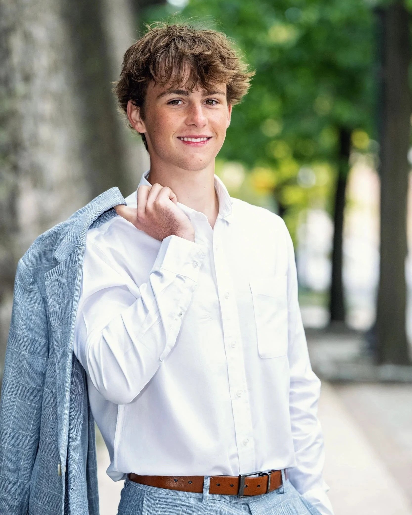 High School senior guy standing in white collared shirt holding suit jacket over shoulder smiling at camera