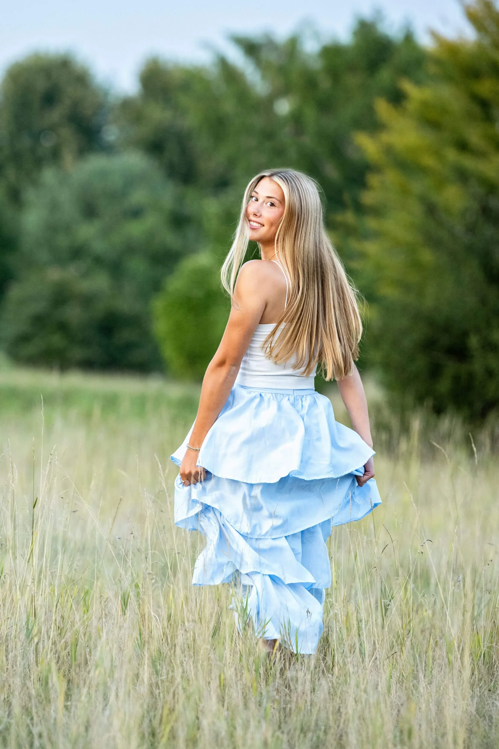 High school senior girl in a blue ruffled shirt and white top running in grassy field looking over shoulder during Tish OConnor Photography portrait session Williamsport PA