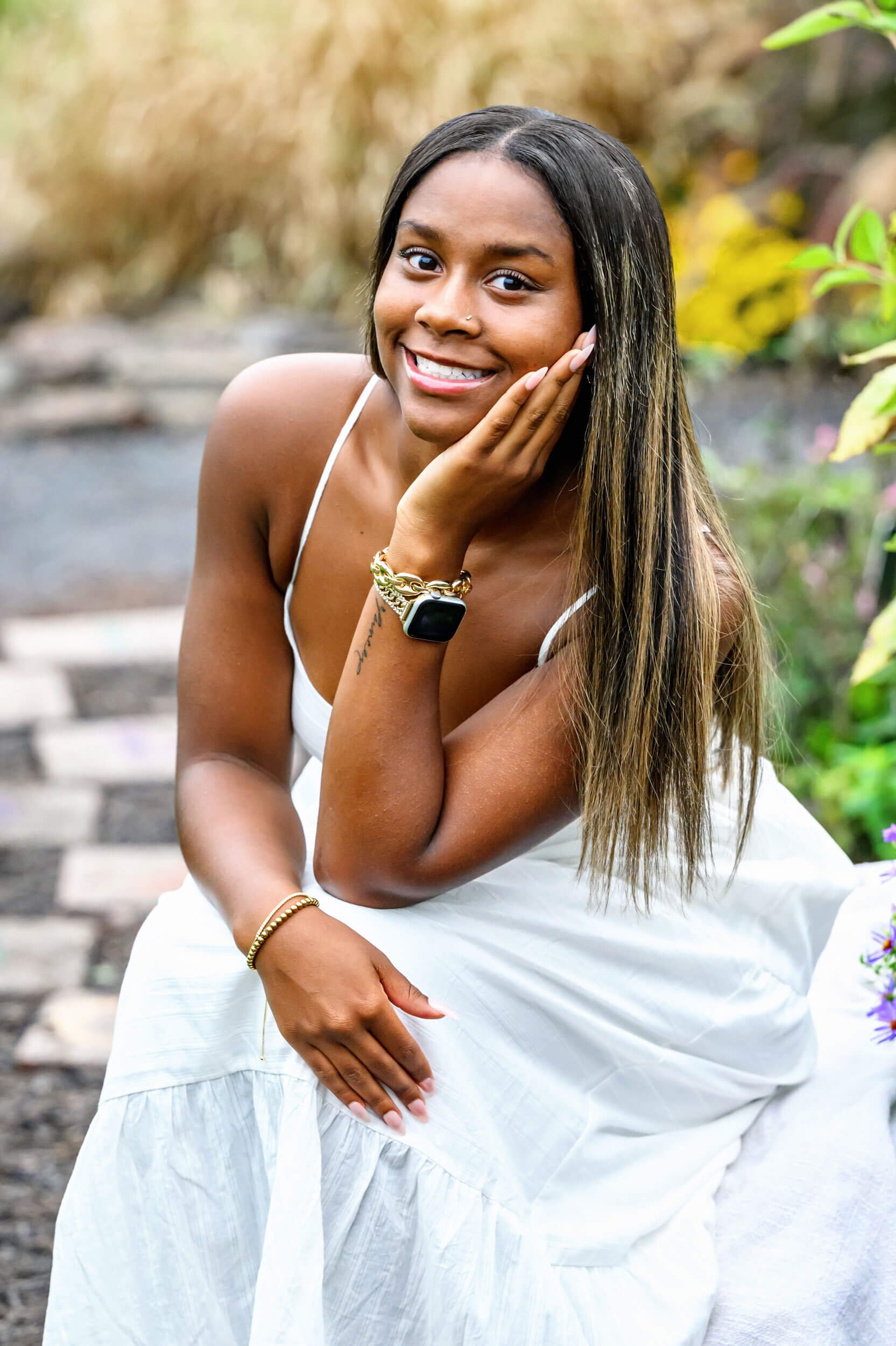 Senior portrait of high school girl in white dress sitting with elbow on knee smiling at the camera in garden setting
