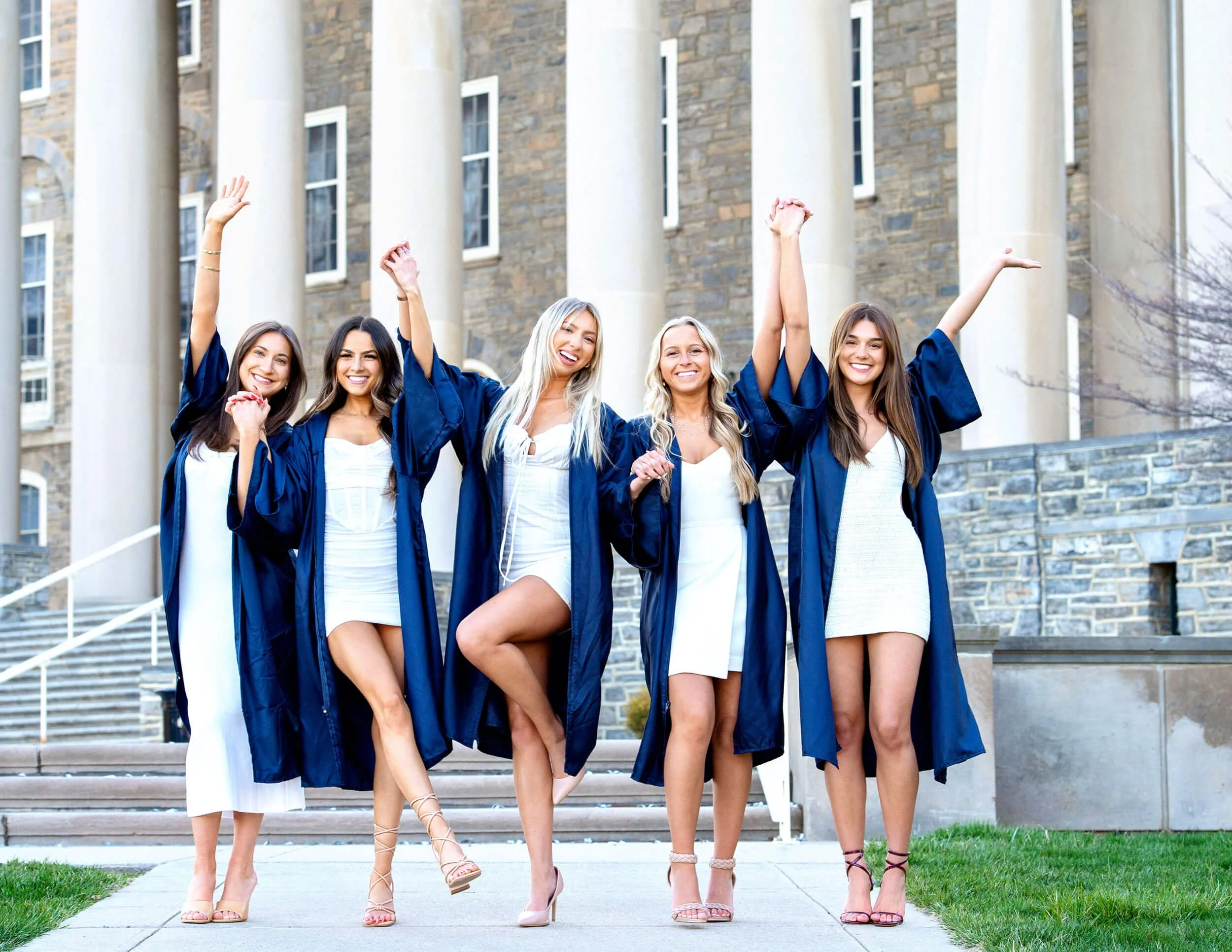 Five female college grads in white dresses with graduation gowns on holding hands and throwing arms in the air in front of Old Main