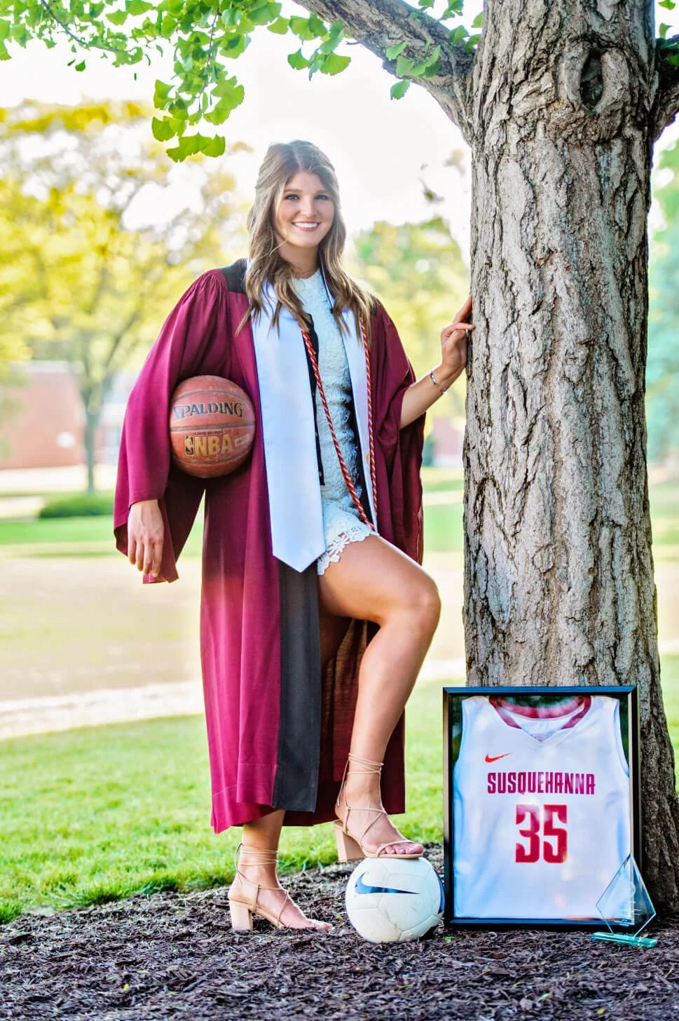 College graduation portrait of Susquehanna University graduate in graduation gown with stole and cords holding a basketball and having a soccer ball under her foot with heels on