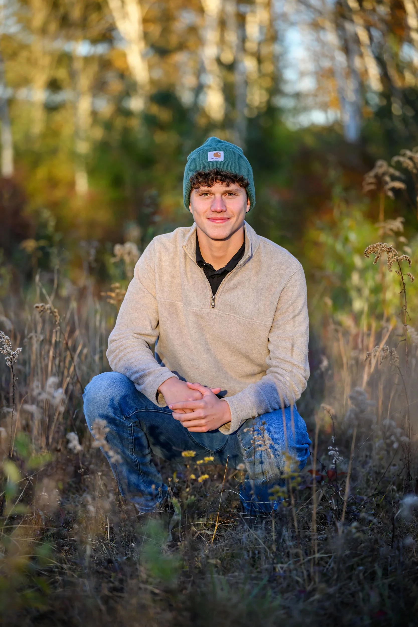 High school senior portrait of a guy in a hat squatting among colorful fall foliage at Rose Valley Lake during an outdoor senior session.