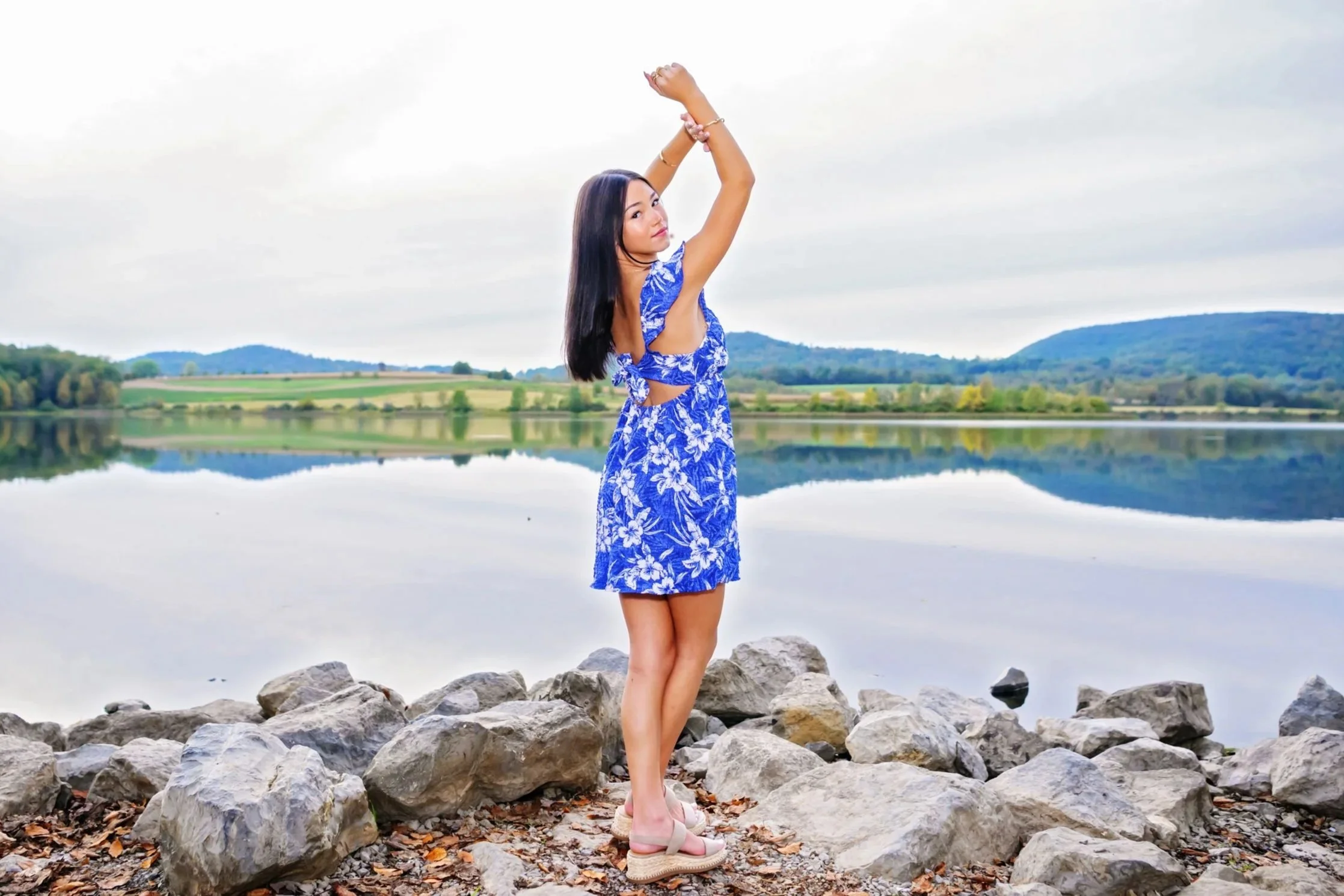 High school senior in a purple dress standing looking over shoulder, arms in the air by a lake portrait session in Williamsport PA