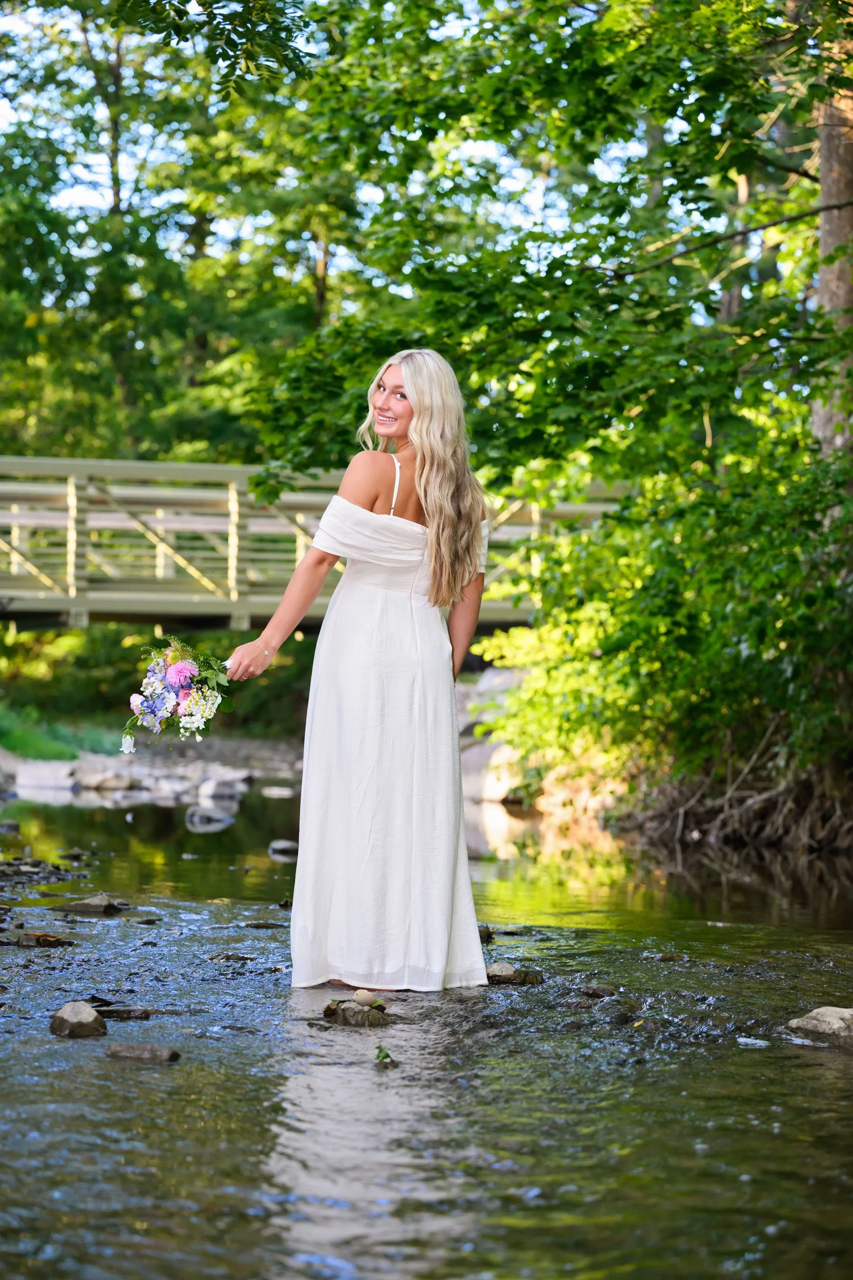High school senior portrait standing in a creek in a long white dress looking over shoulder for senior session in Williamsport, Pennsylvania.