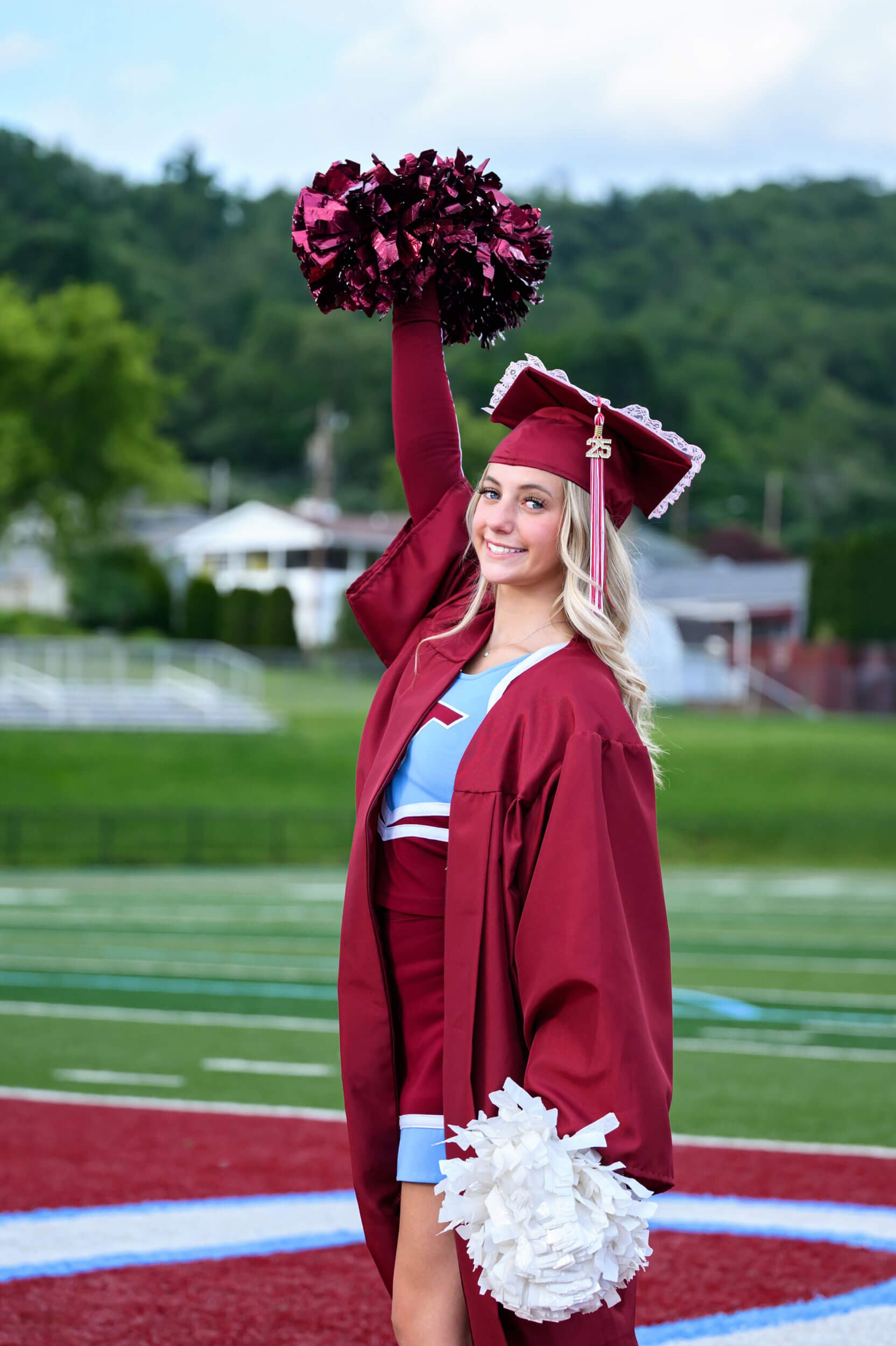 High school graduation senior portrait of chearleader in uniform with cap and gown on standing on football field with pom poms 
