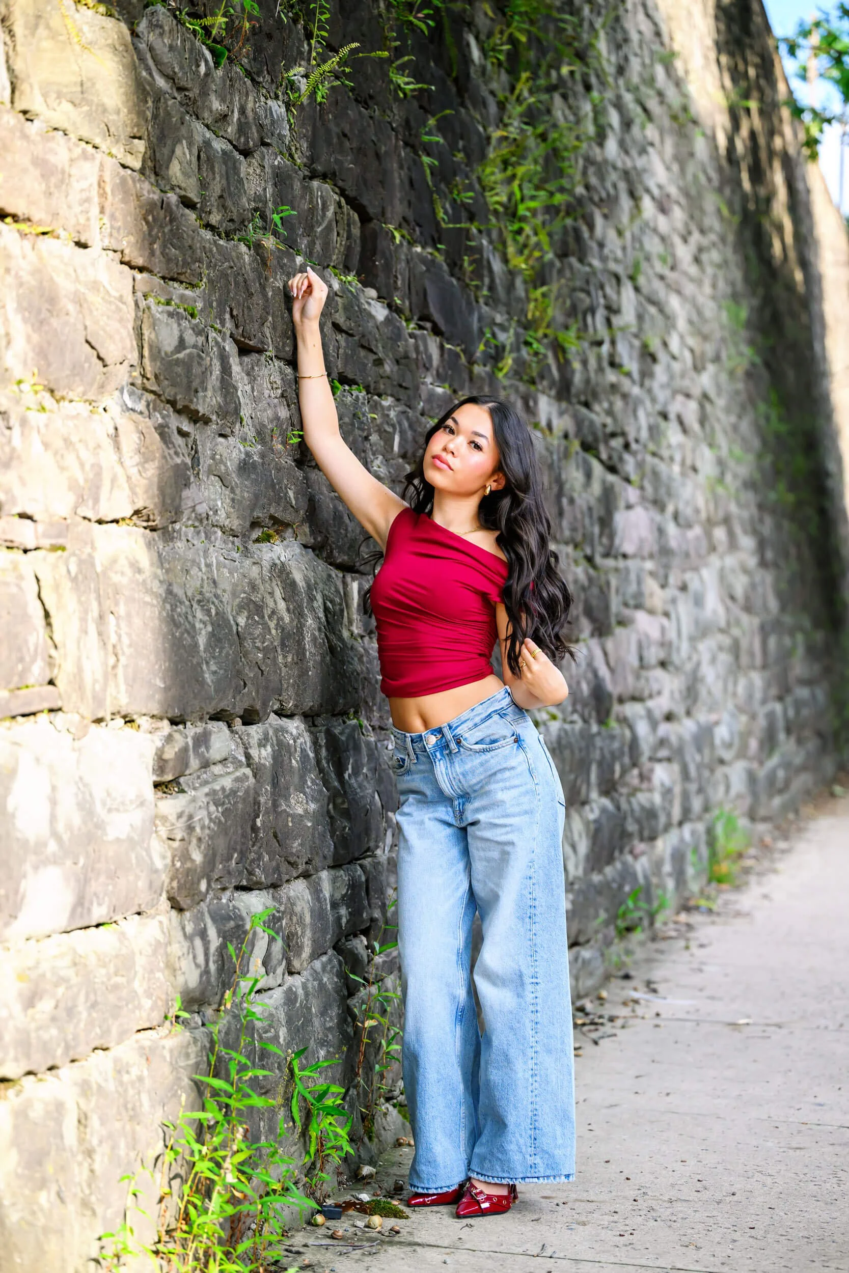 Tish OConnor Photography senior portrait of girl next to stone wall in downtown Williamsport with red top, blue jeans and red heels with arm up on wall