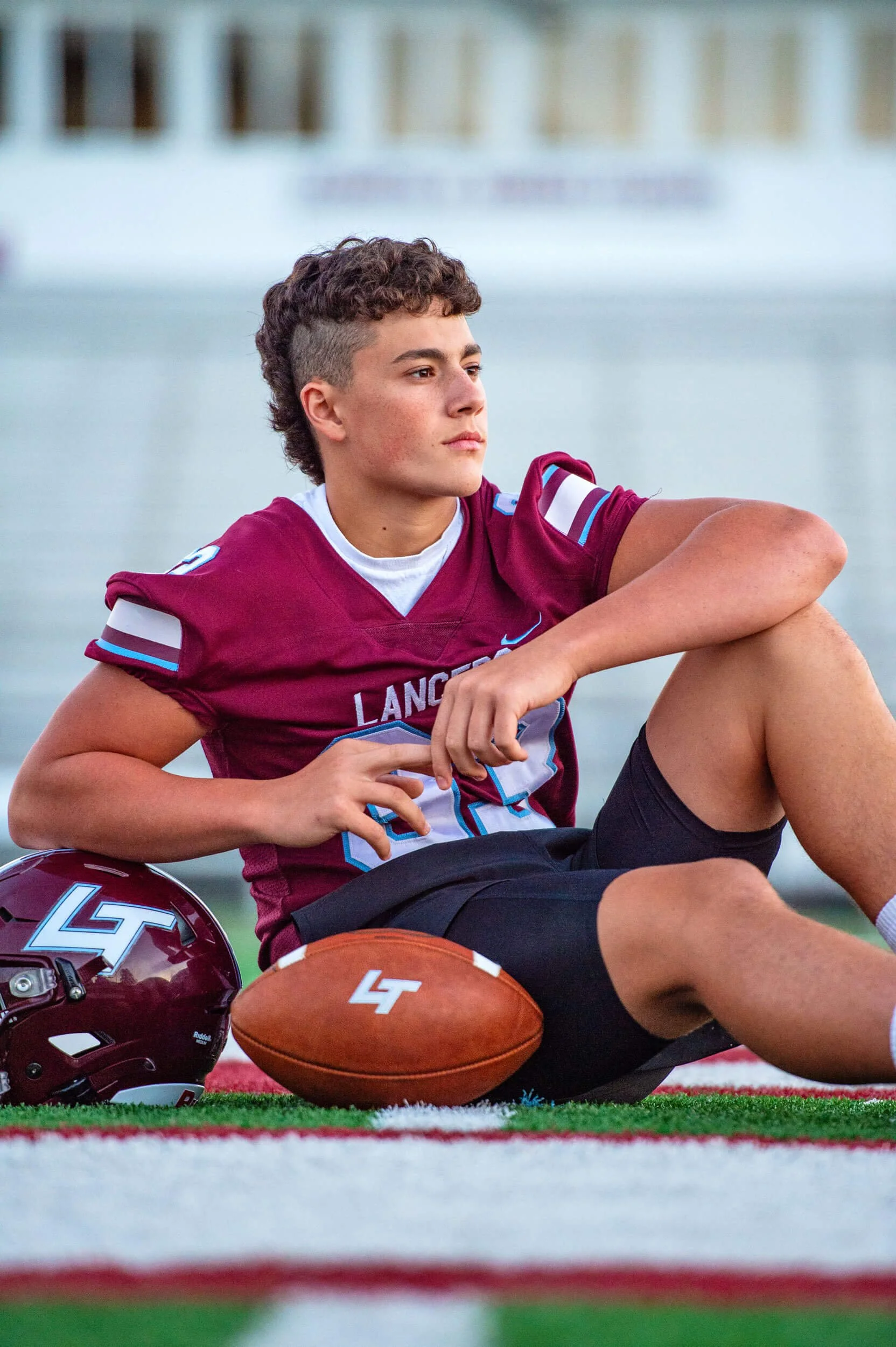 Loyalsock Township High School senior guy sitting in football jersey on the football field leaning his elbow on his helmet with football in front 