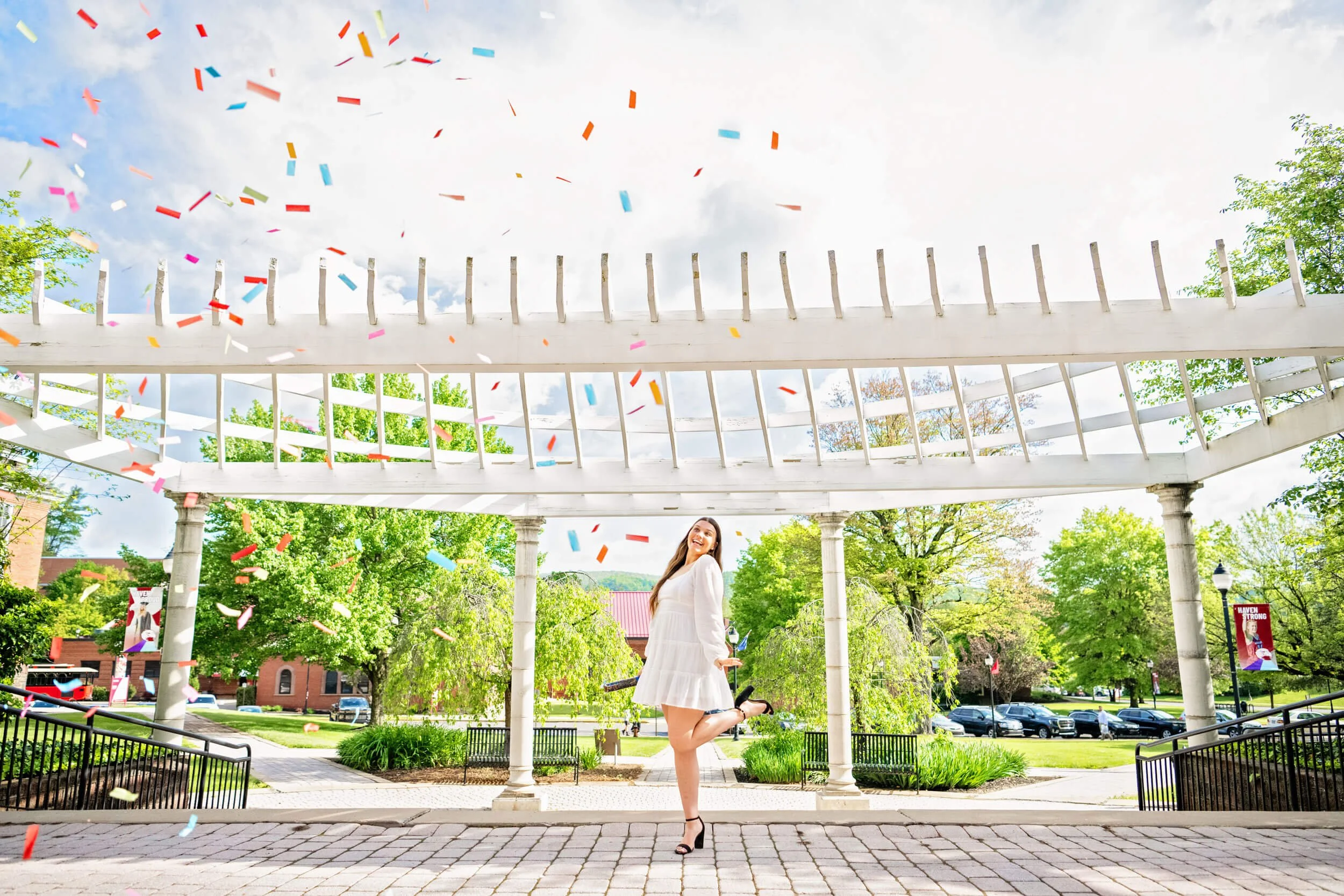 College graduation portrait of Lock Haven University grad in white dress standing under a white pergola on campus shooting confetti into the air 