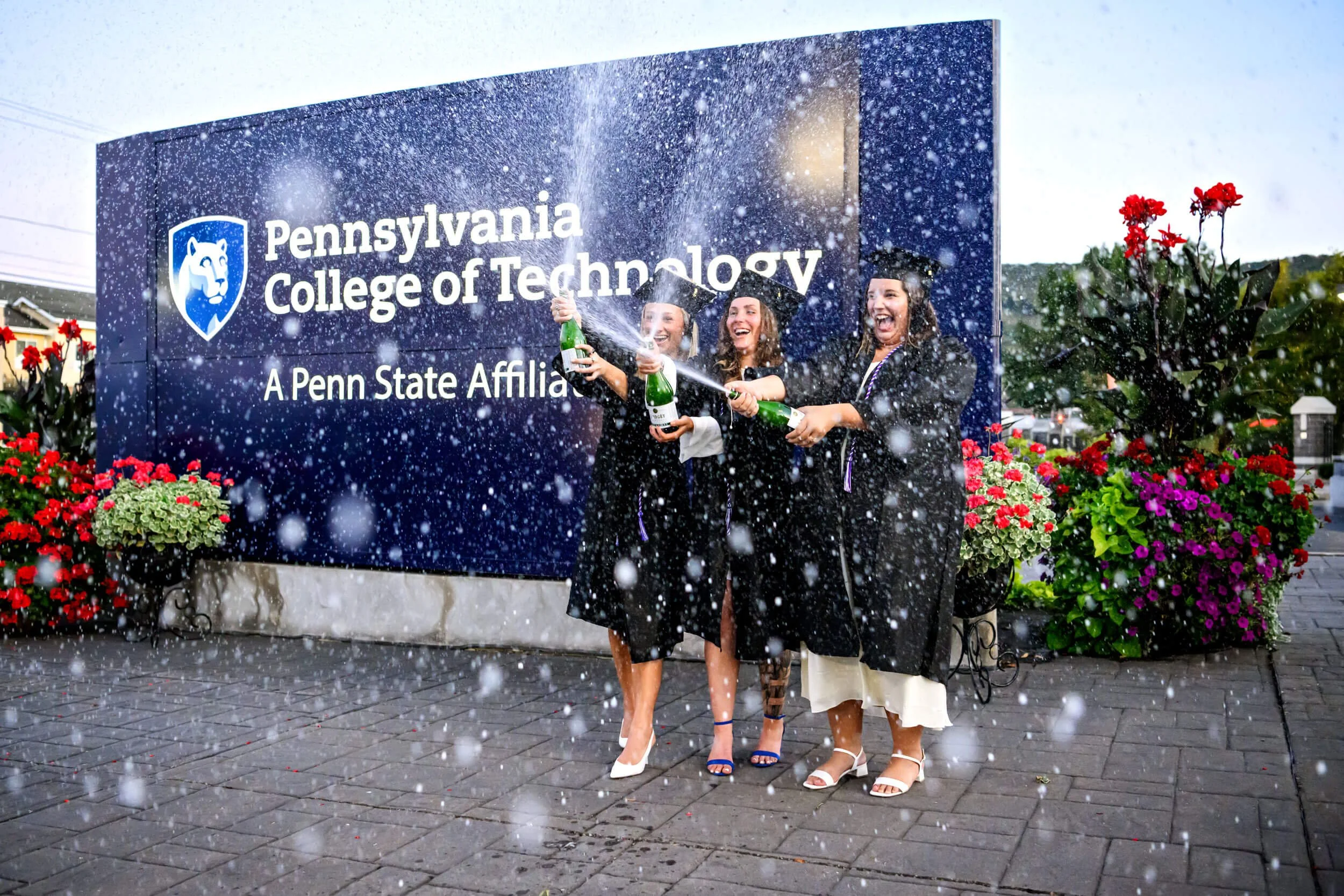 Three Penn College grad girls in caps and gowns spraying champagne by the Penn College of Technology sign grad photos