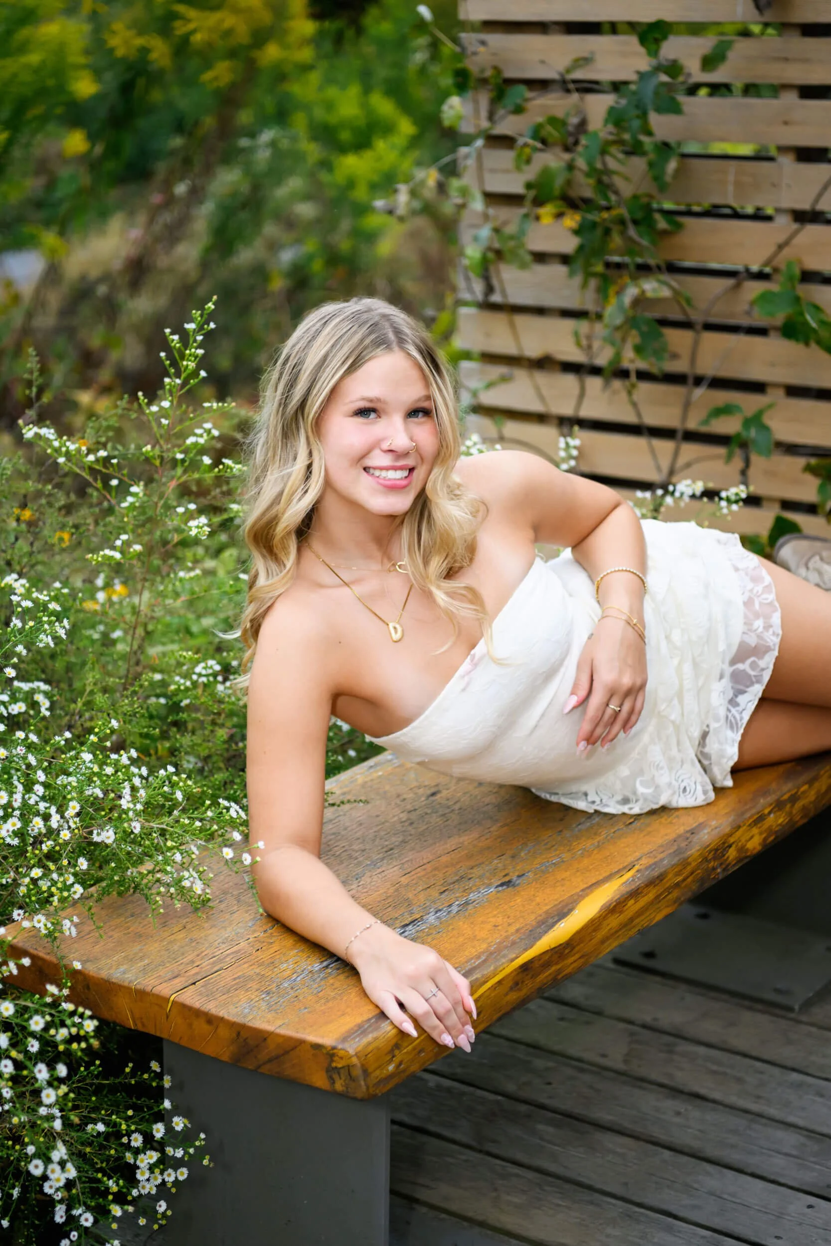 girl in short white lacy dress laying on wooden bench with greenery around her for senior portrait session with williamsport senior photographer Tish OConnor 